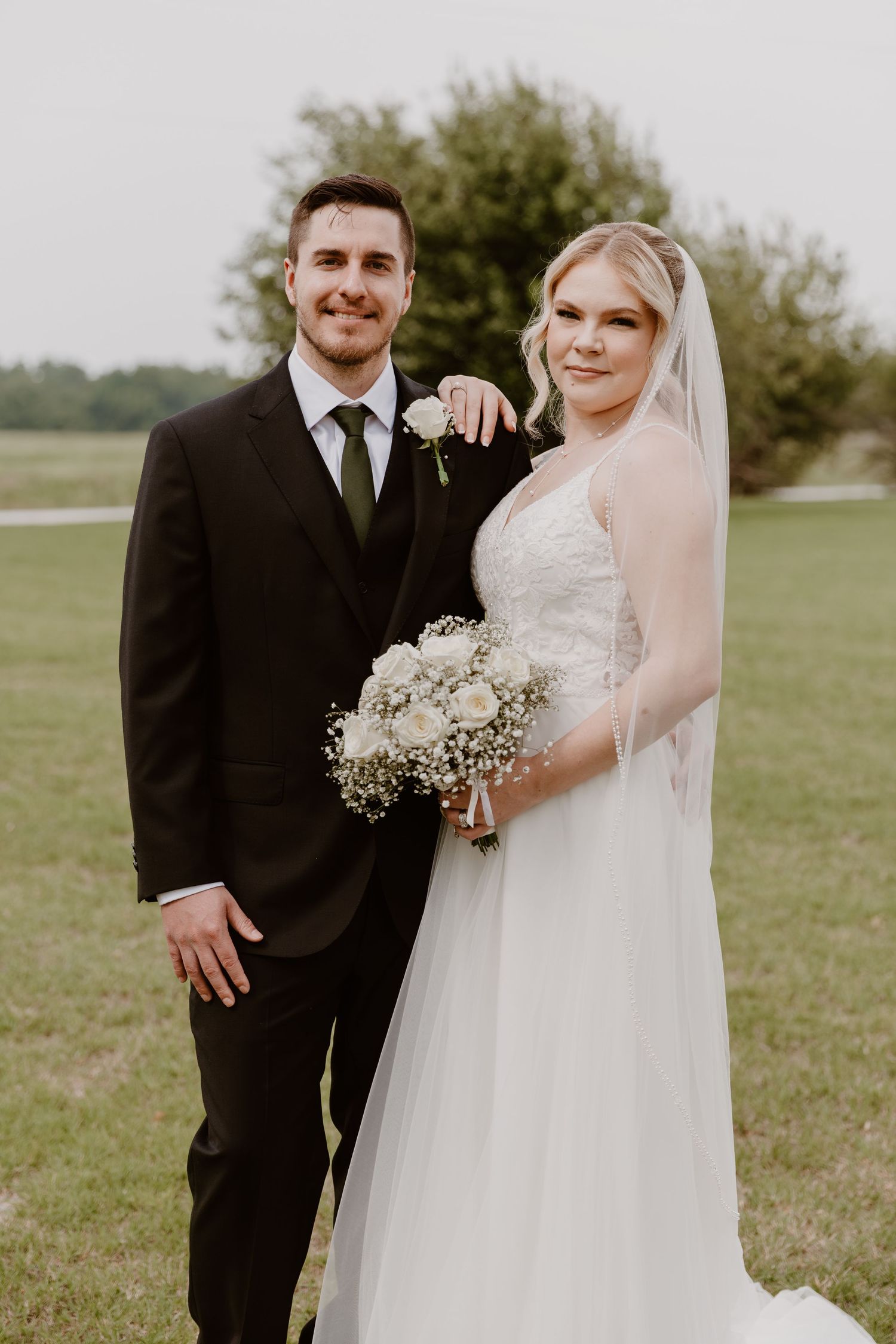 Newlywed couple poses outdoors, bride in white gown with veil and groom in black suit with boutonniere.