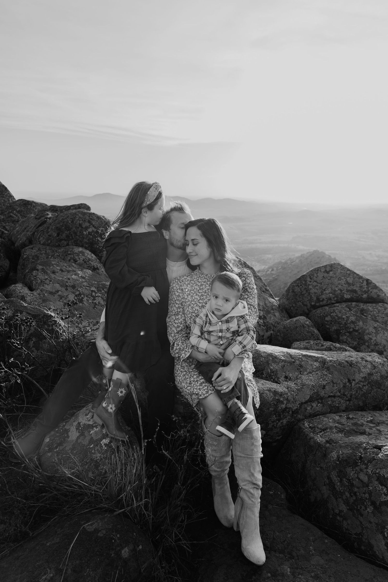A couple sits together on rocky cliffs overlooking a scenic mountain landscape in black and white photography.