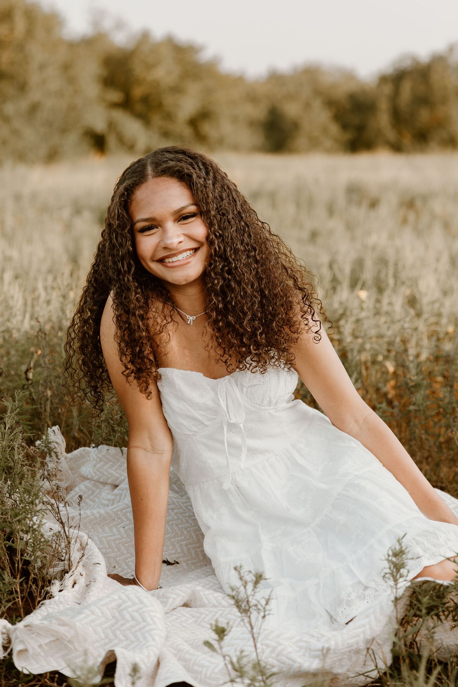 A joyful portrait of someone with curly hair in a white dress sitting in a sunlit meadow.