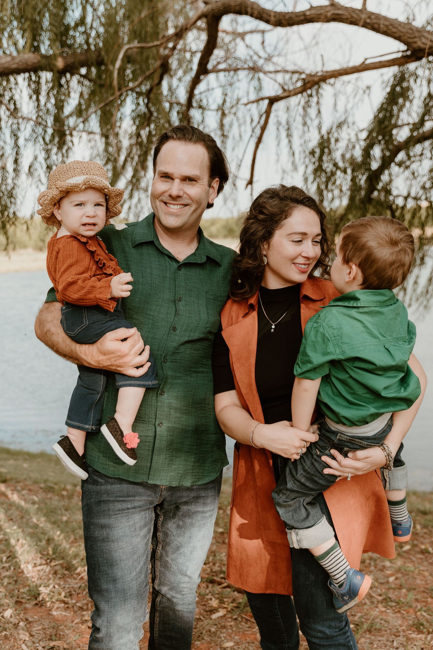 Family wearing green and rust-colored clothing poses together by a lake during autumn.