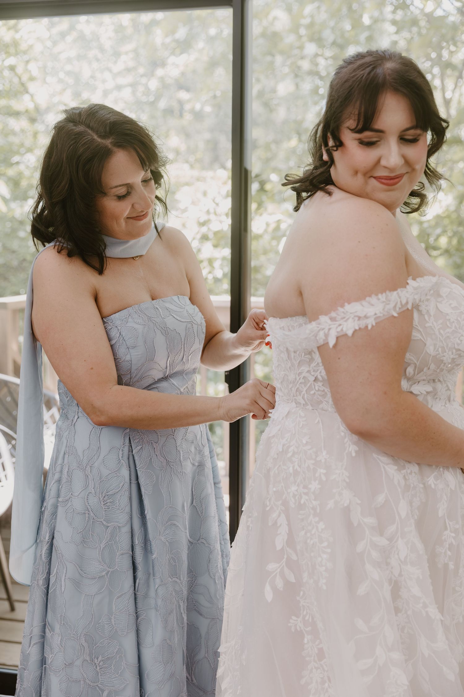 Bridesmaid in blue dress helping bride fasten her off-shoulder lace wedding gown before the ceremony.