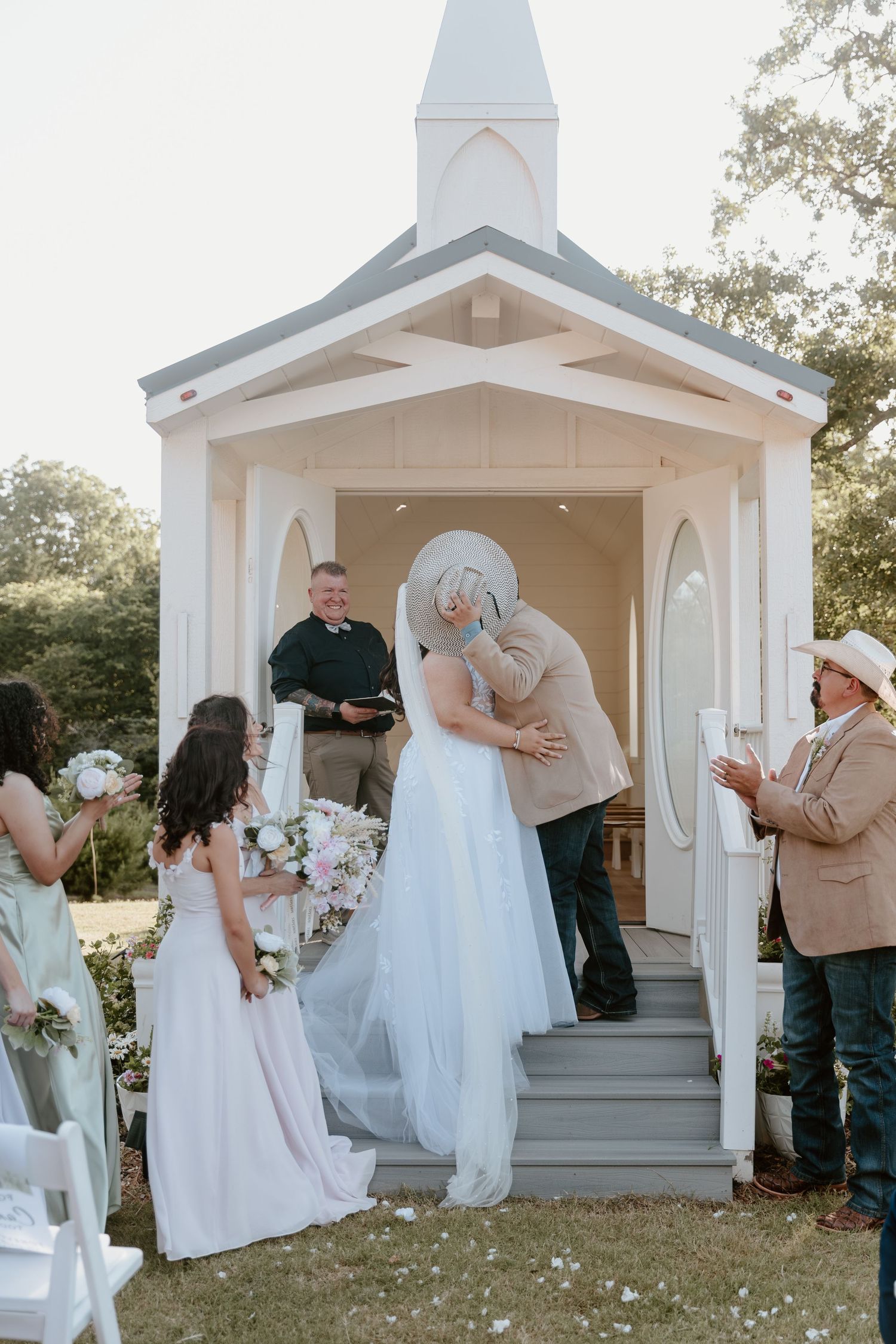 Bride and groom share first kiss on steps of white outdoor chapel during romantic garden wedding ceremony.