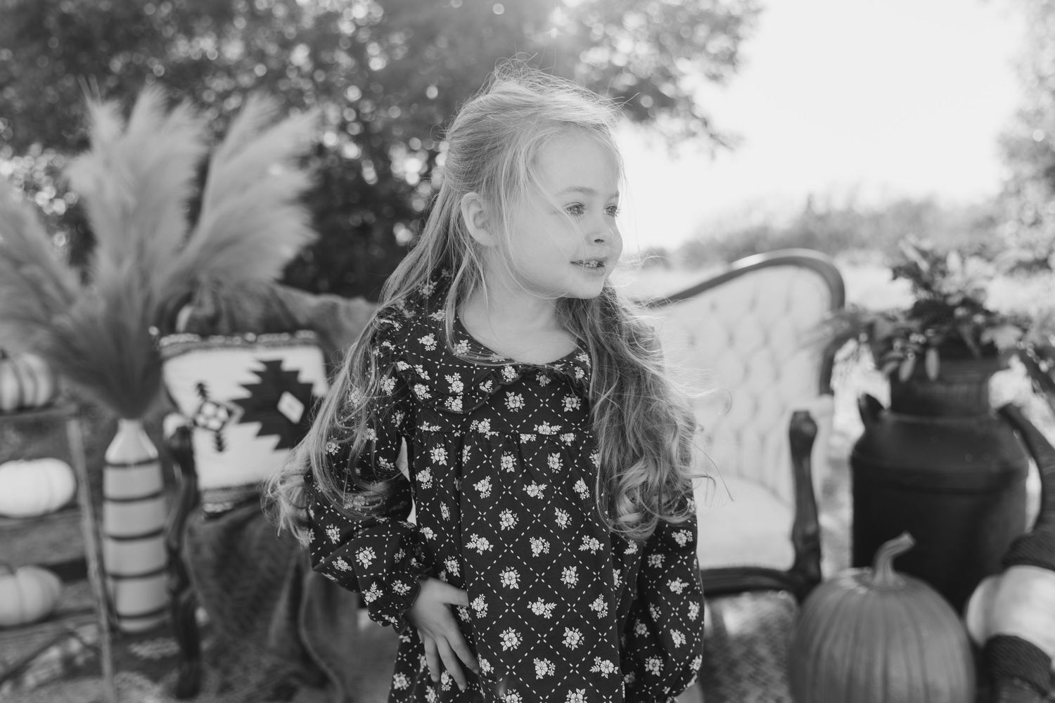 A black and white photograph of a child in a floral dress standing outdoors near pumpkins.