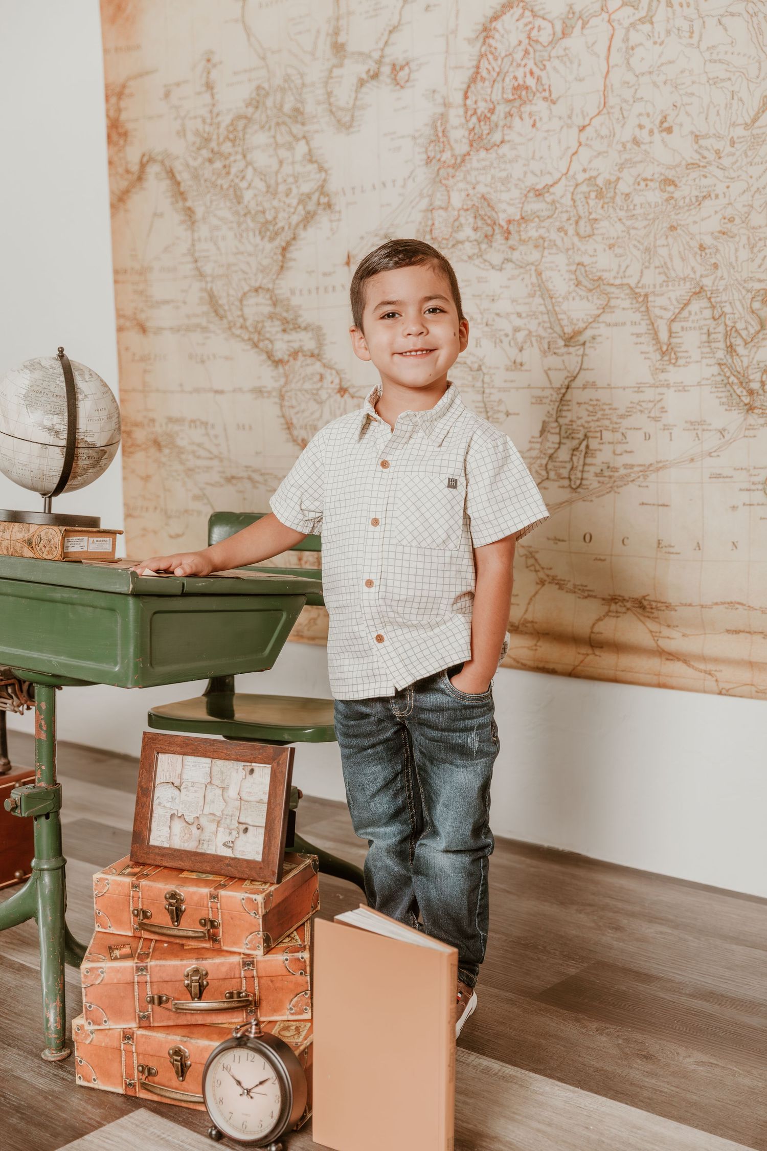 A young boy stands next to a green desk and vintage suitcases with a world map background in a rustic-styled setting.