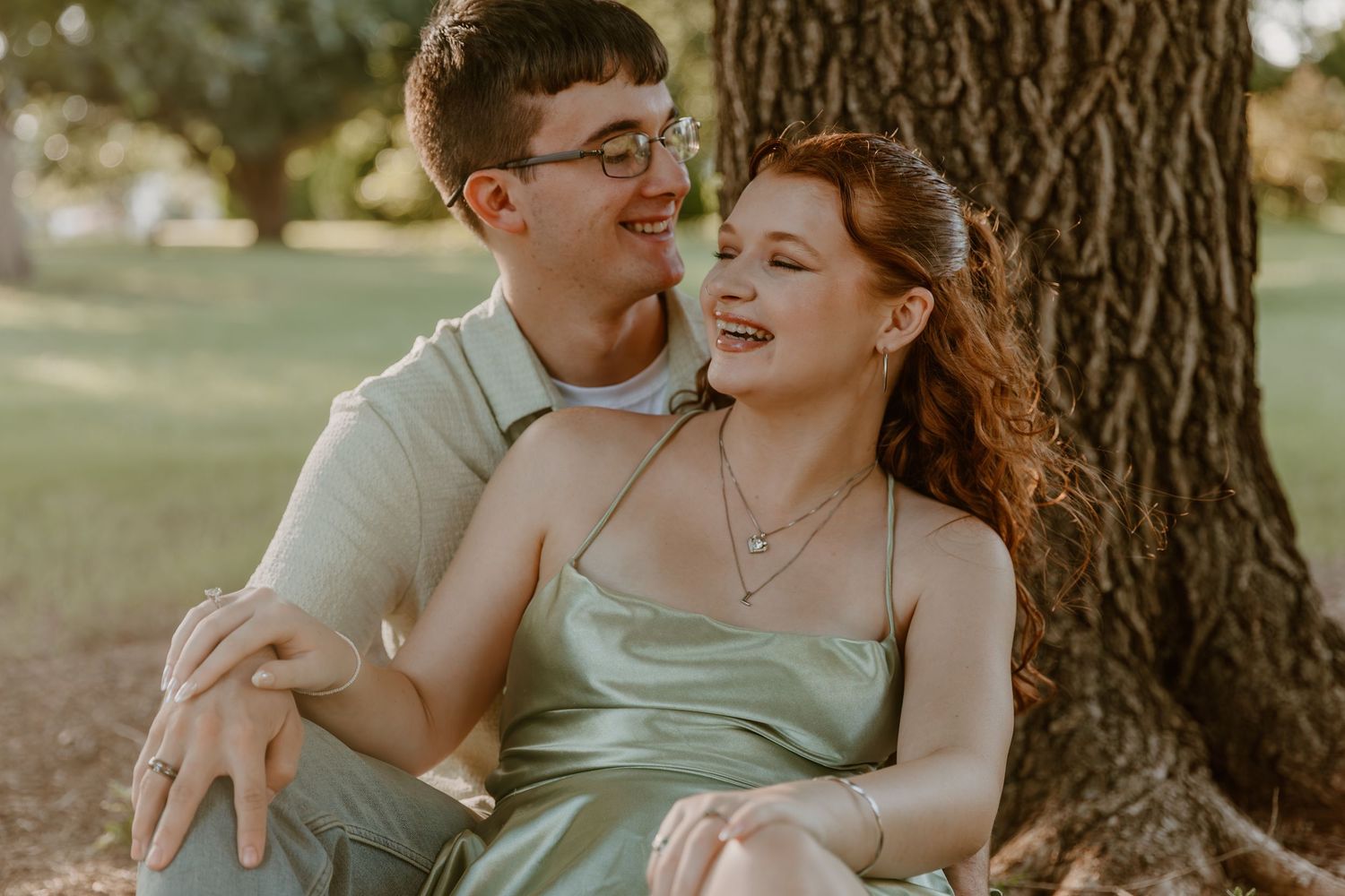 A couple shares a joyful moment together while sitting under a large tree in warm sunlight.