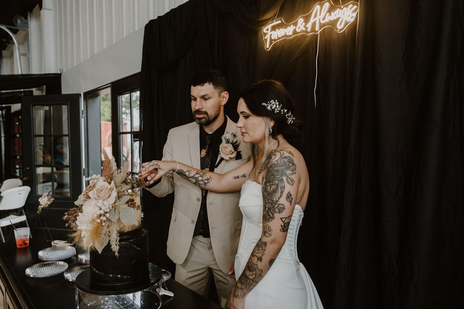 Tattooed bride and groom cutting black wedding cake under Forever & Always neon sign at elegant reception.