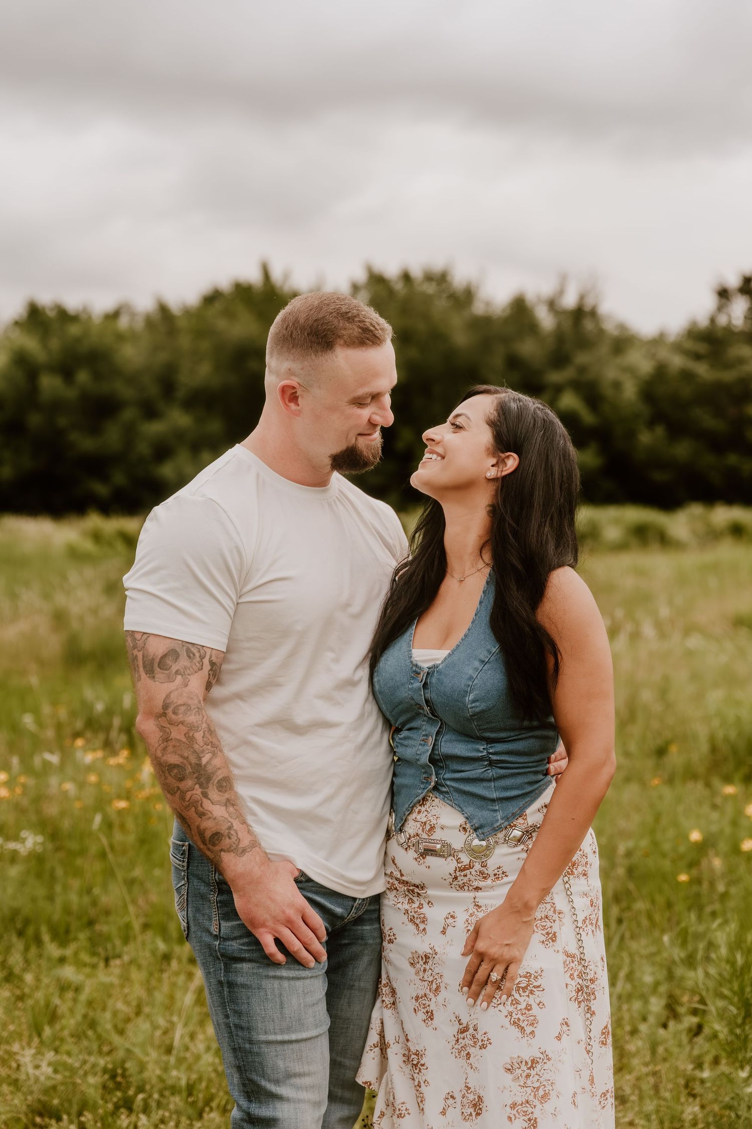 A couple shares a romantic moment in a green field, the woman in a denim top smiling up at the tattooed man in a white tee.