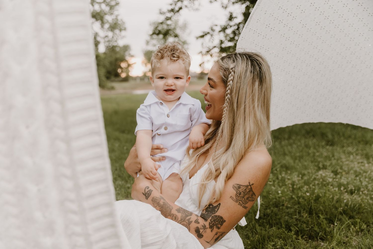 A tattooed blonde mother smiles lovingly at her happy baby boy outdoors in a soft, dreamy natural setting.