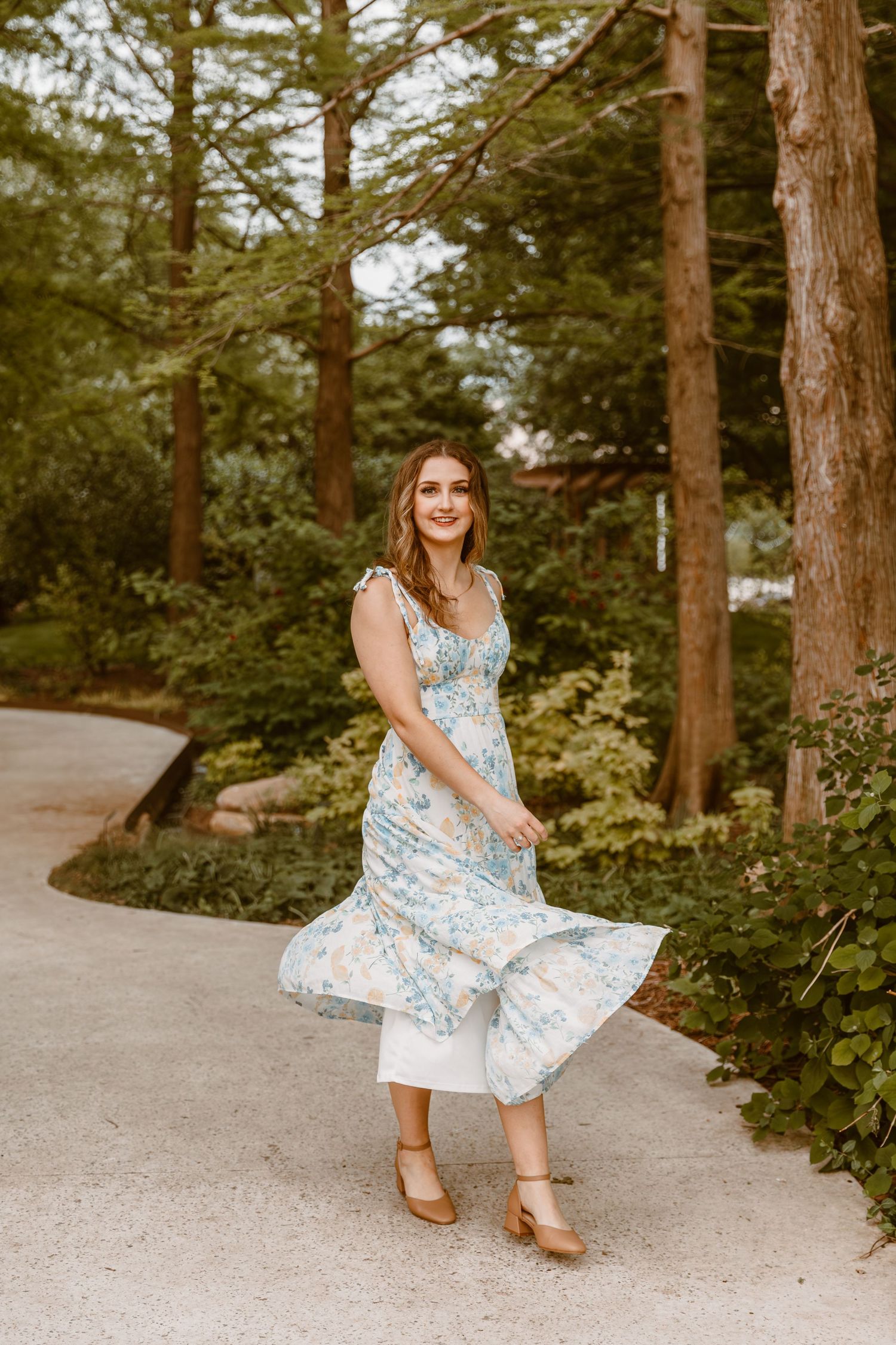 A flowy blue and white floral dress sways on a winding garden path surrounded by tall evergreen trees.