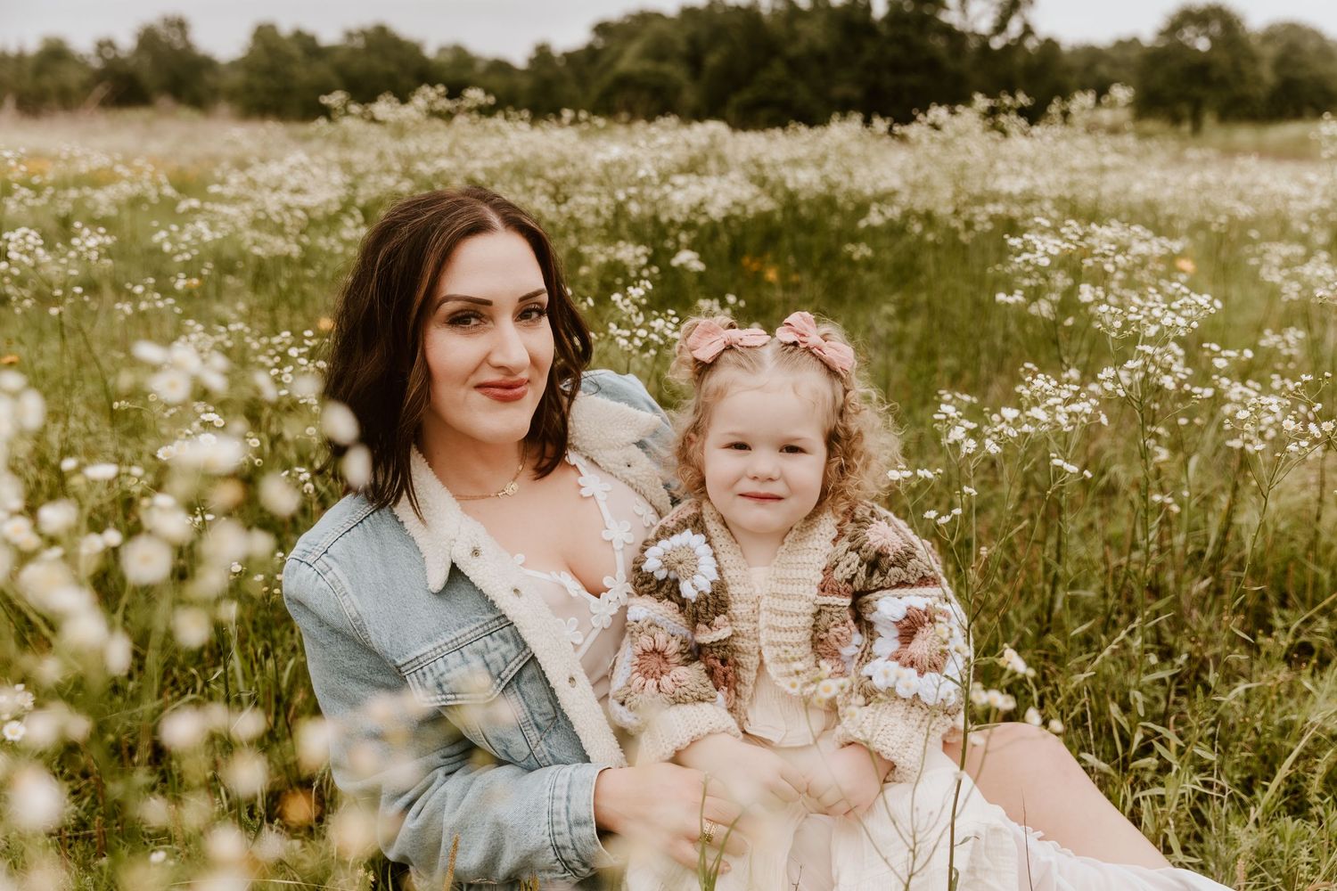 A mother and toddler girl sit together in a blooming wildflower field, both wearing floral outfits, smiling warmly.
