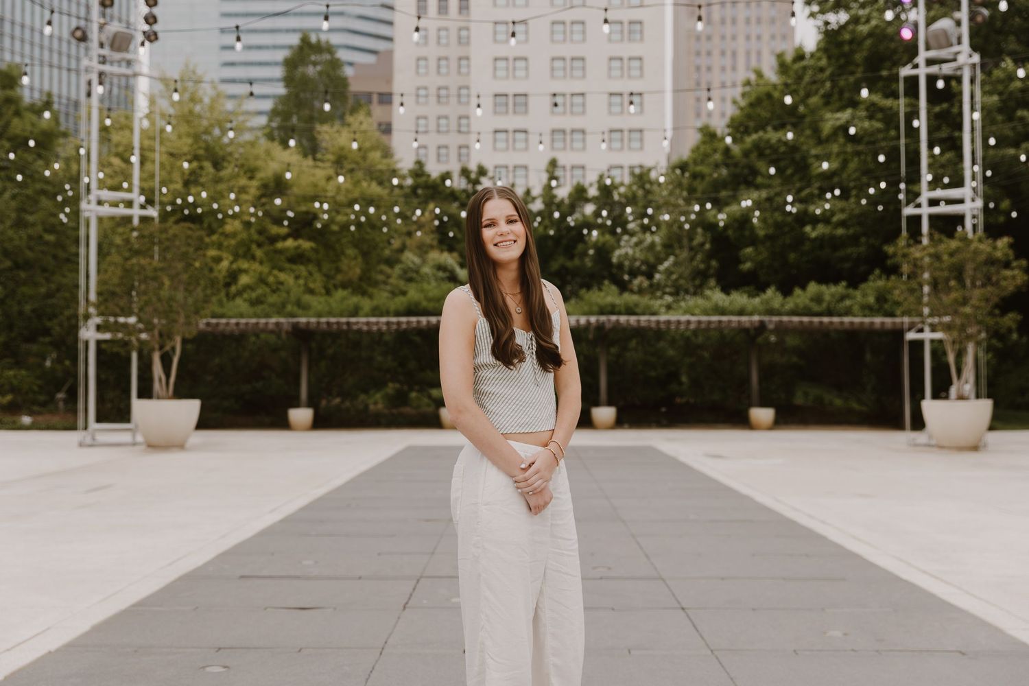 Young woman in white outfit smiling outdoors at urban plaza with string lights and city buildings in background.