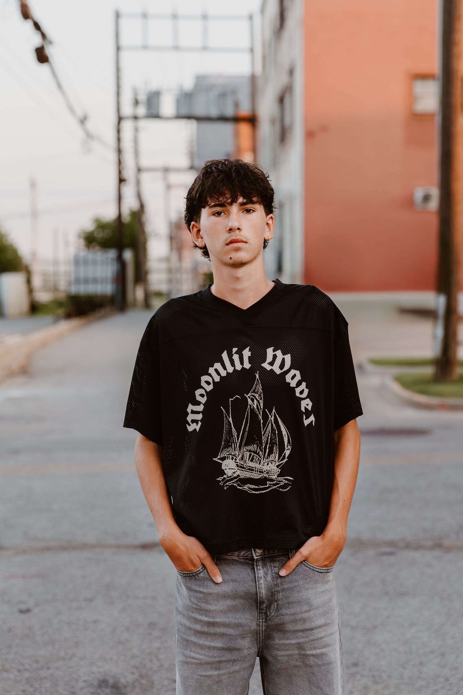 A person wearing a black graphic t-shirt with a ship design and gray jeans stands against an urban backdrop.