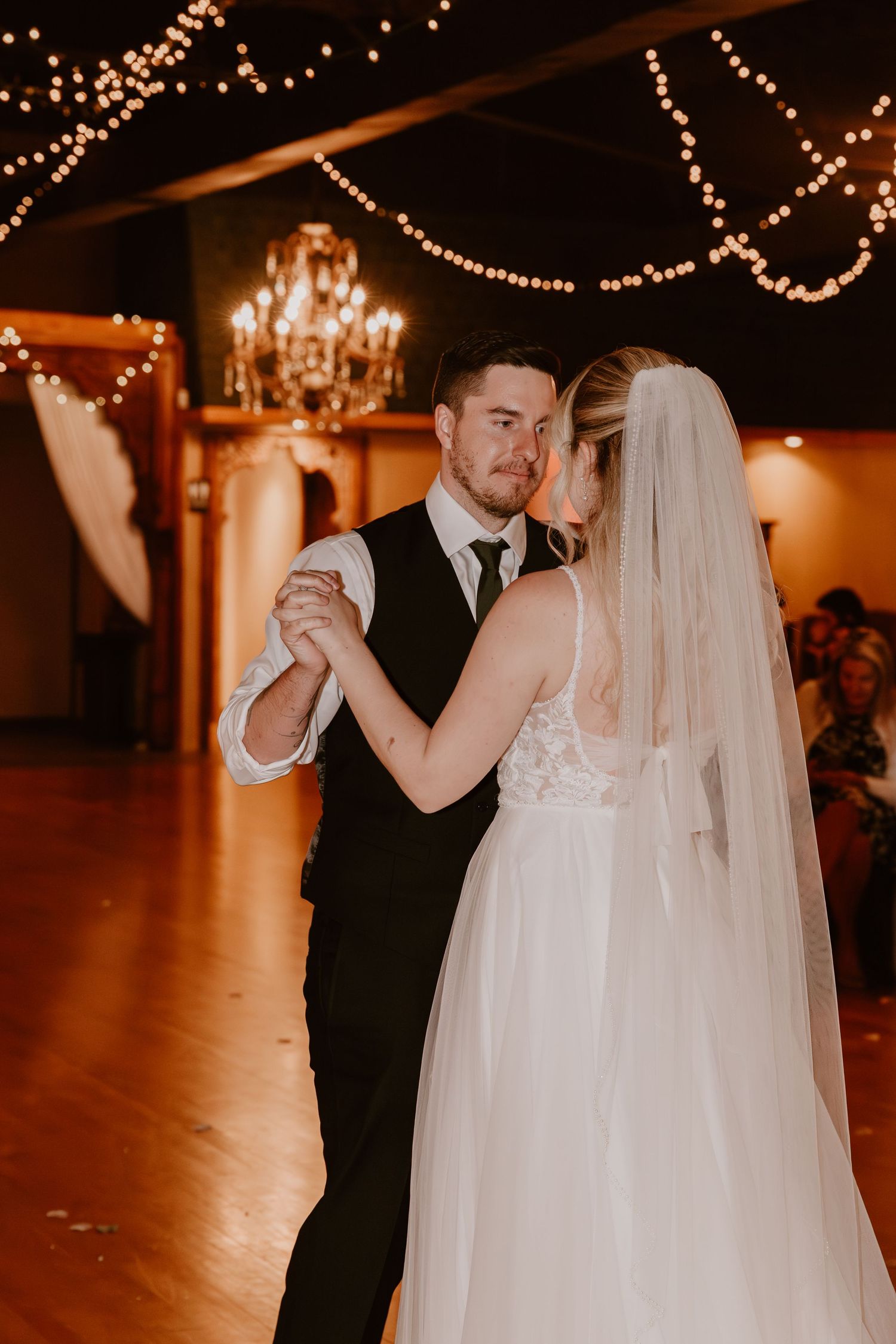 Newlyweds dance together in an elegant ballroom decorated with romantic string lights and warm ambient lighting.