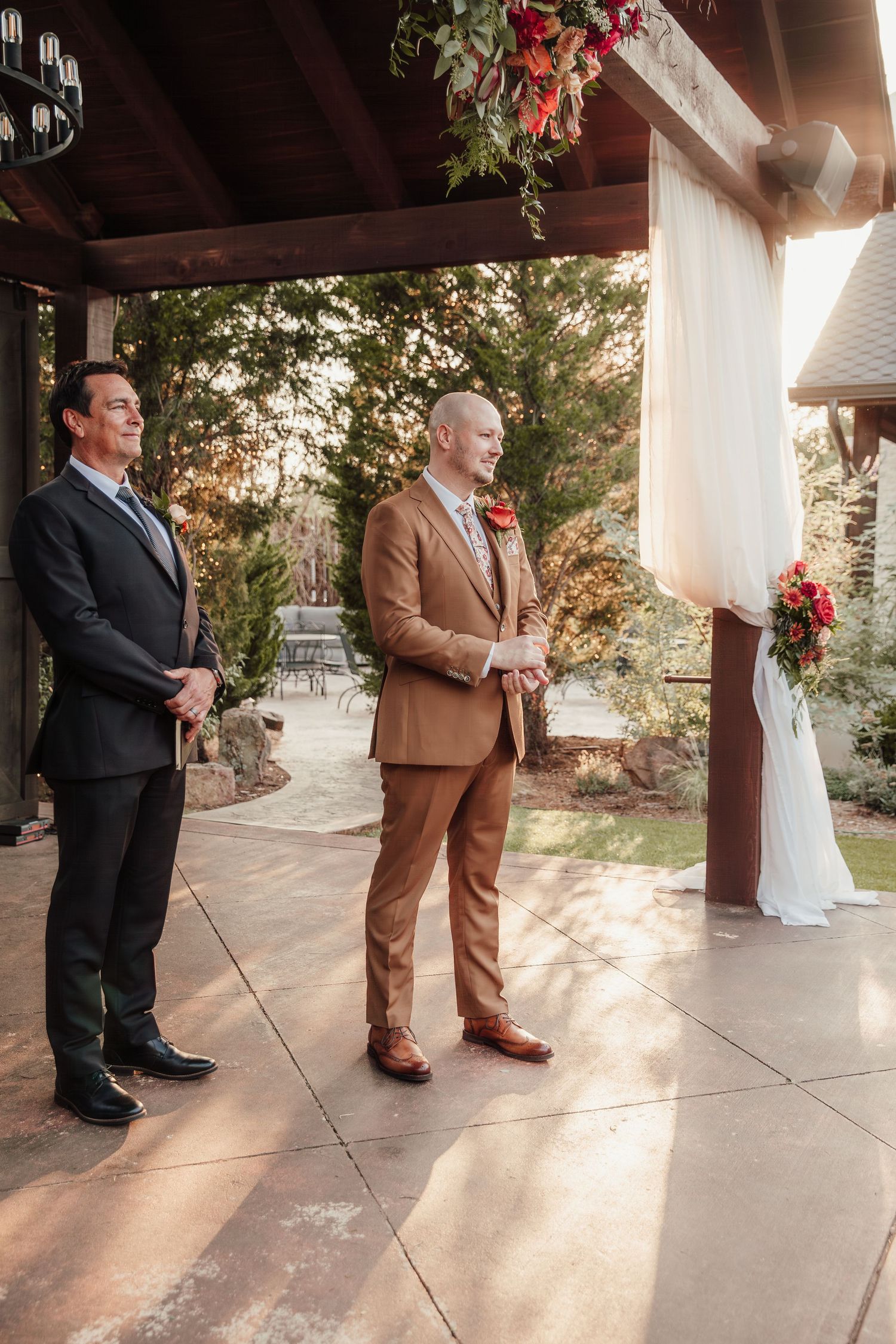 Two men stand under an outdoor wedding pavilion decorated with floral arrangements and white draping in warm sunlight.