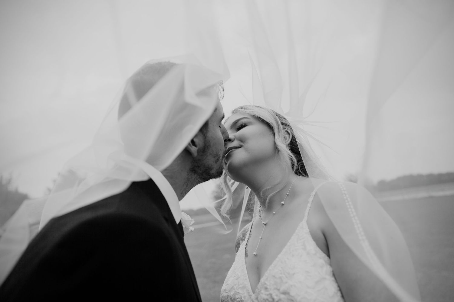 A couple shares a romantic kiss under a flowing veil during their black and white wedding photograph.