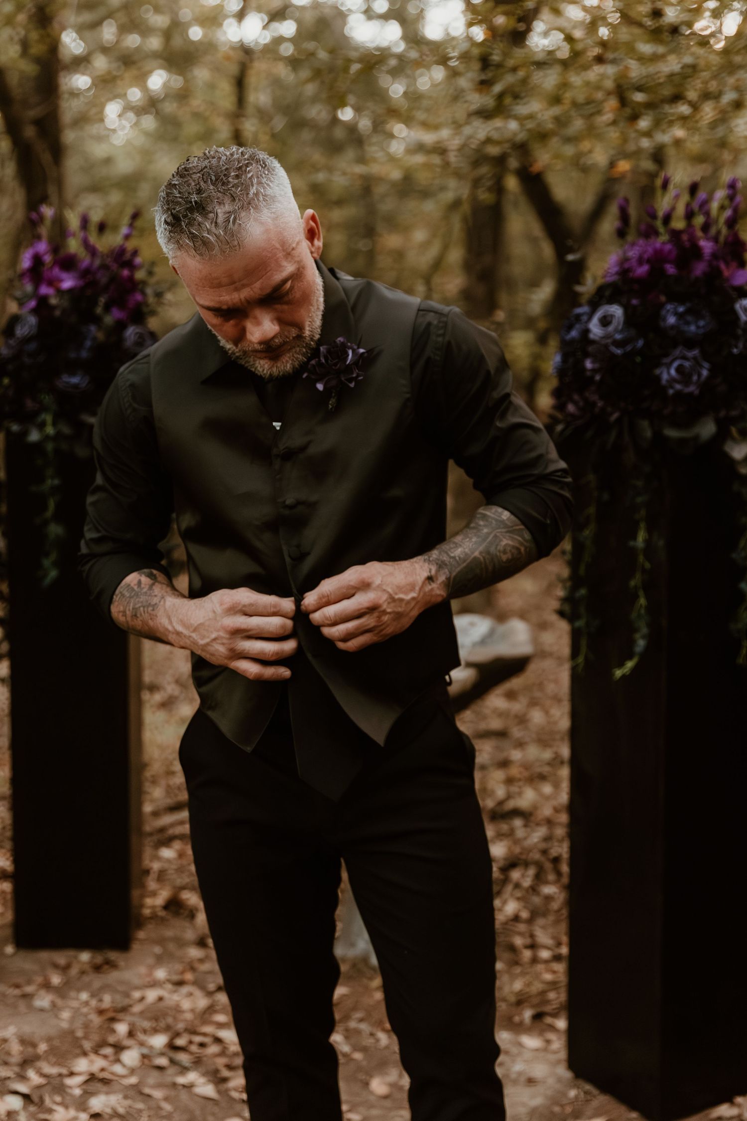 A person in black attire examines something closely while standing in a wooded area with purple flowers in the background.