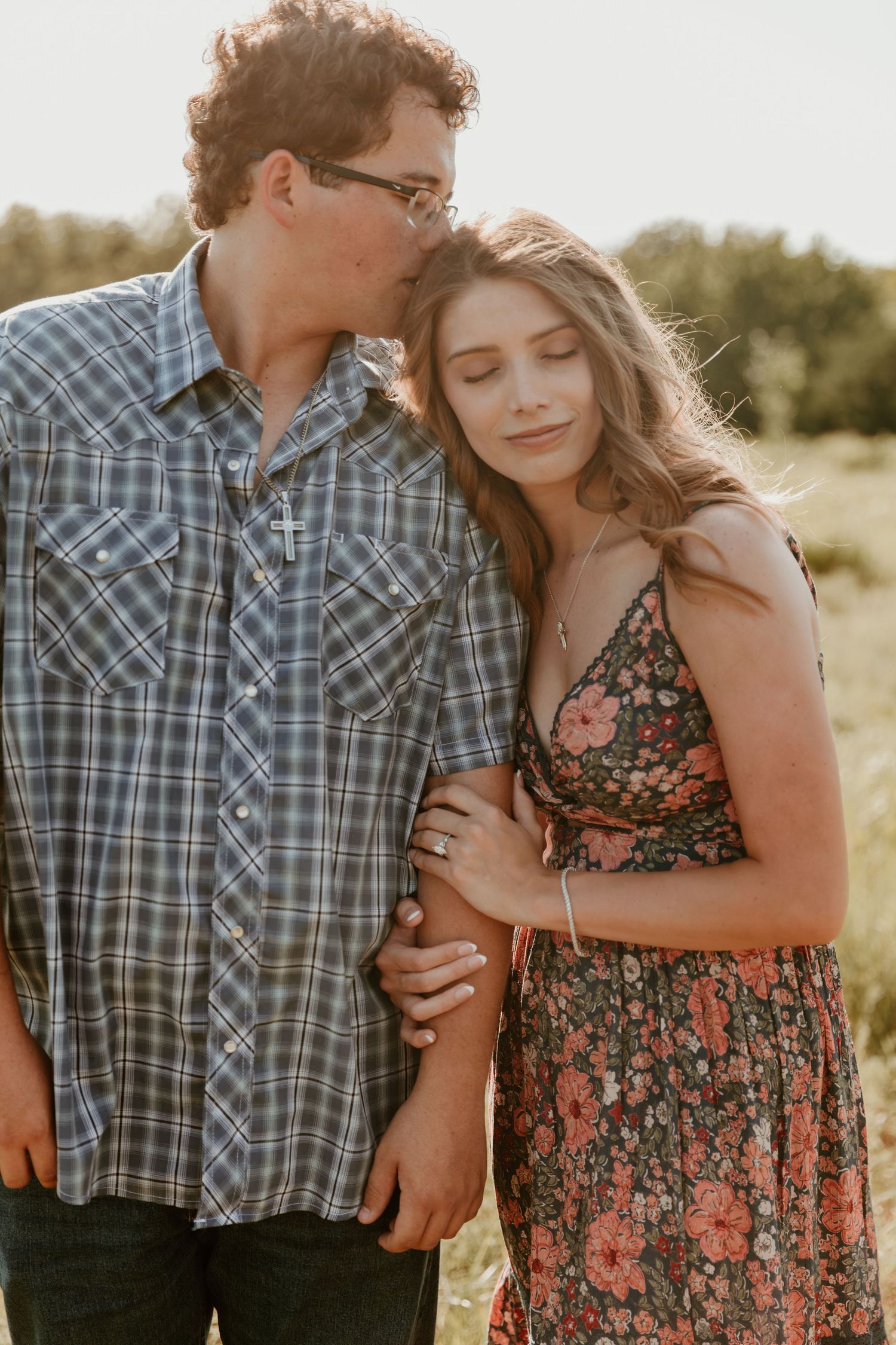 A couple embraces in a sunset field, wearing plaid and floral patterns while sharing an intimate moment.