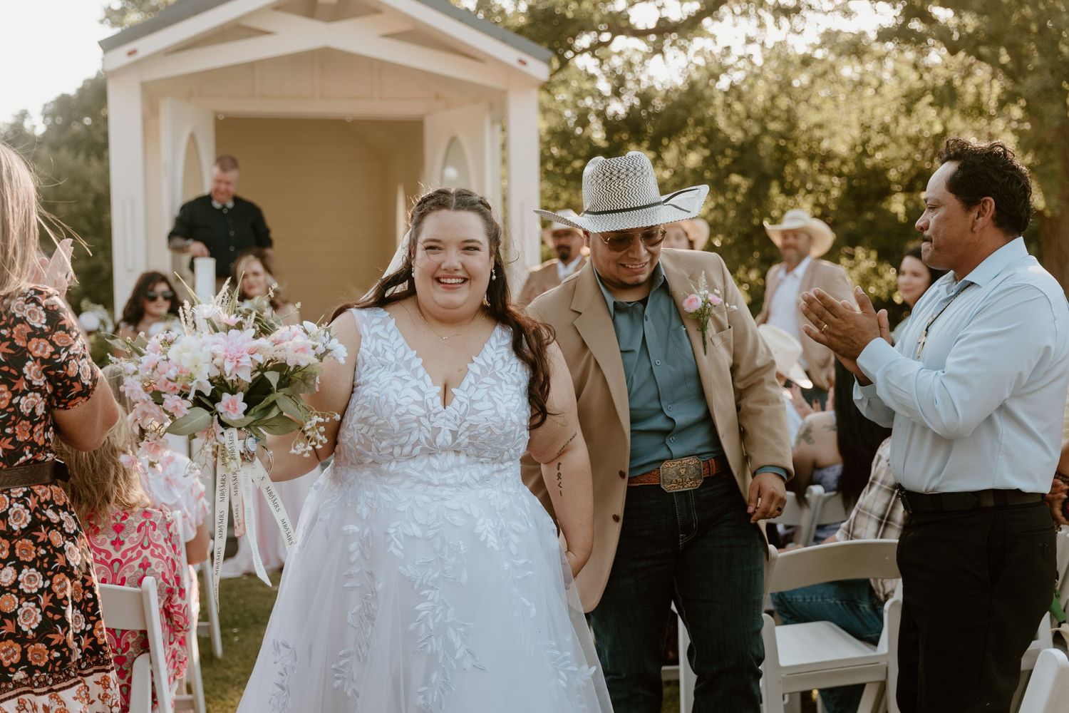 Bride in white lace gown walks down outdoor aisle with man in cowboy hat, guests applauding at rustic wedding.