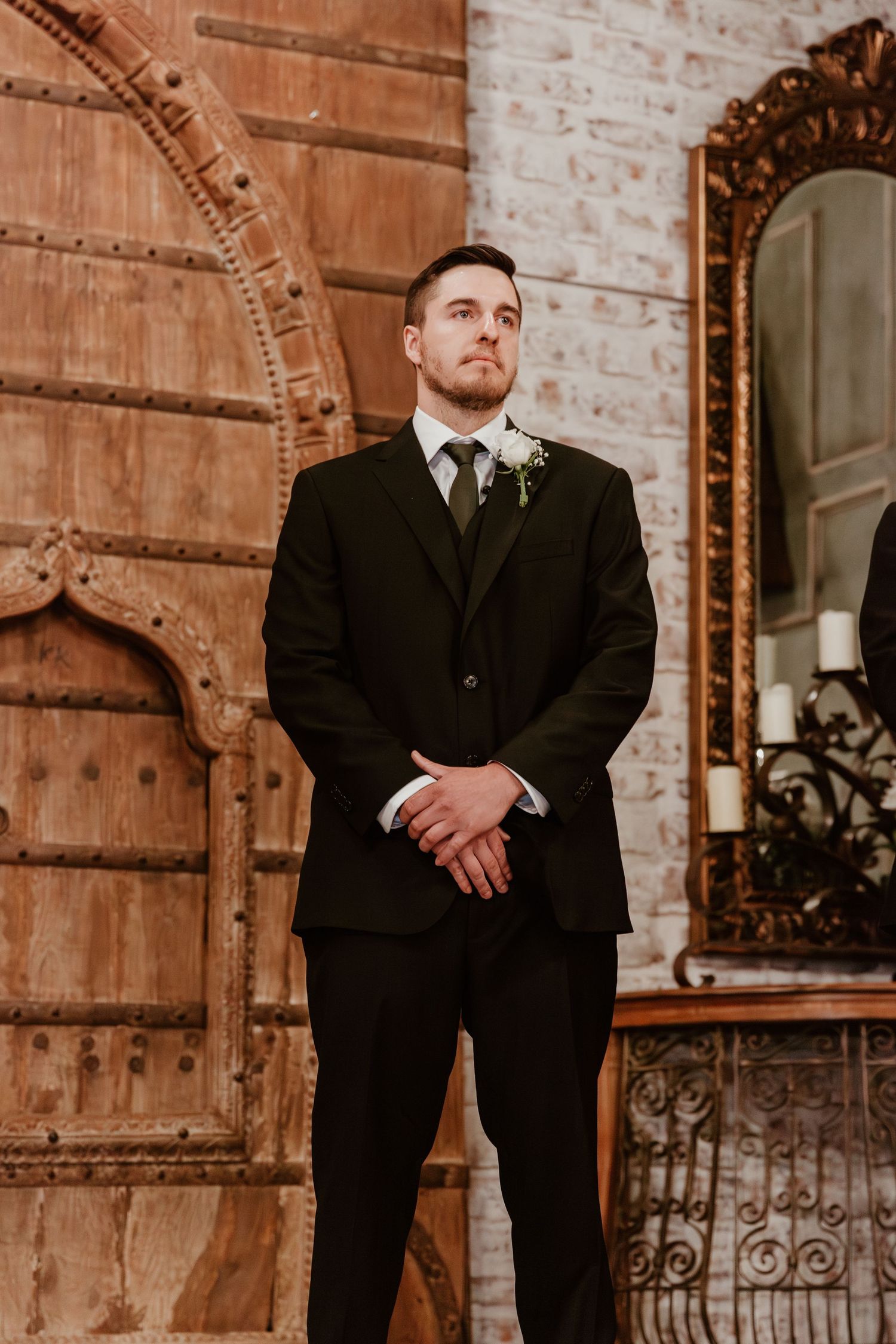 A groom in a black suit stands formally against a brick wall with an ornate mirror and archway.