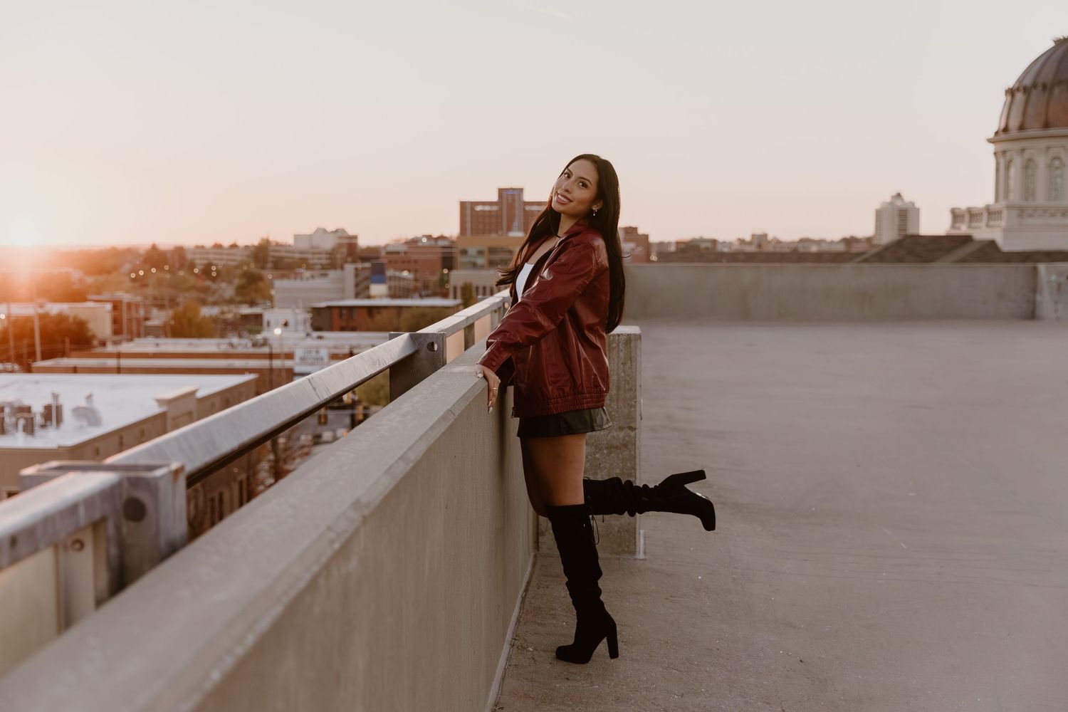 Person in a brown coat stands on a rooftop at sunset overlooking a cityscape in warm golden light.
