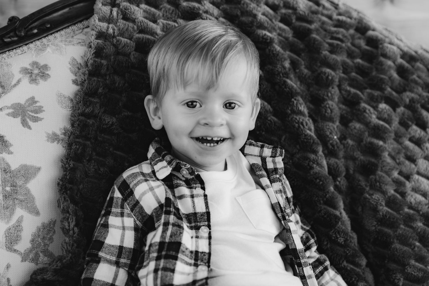 A joyful young child wearing a plaid button-up shirt over white sits on a textured couch in black and white.
