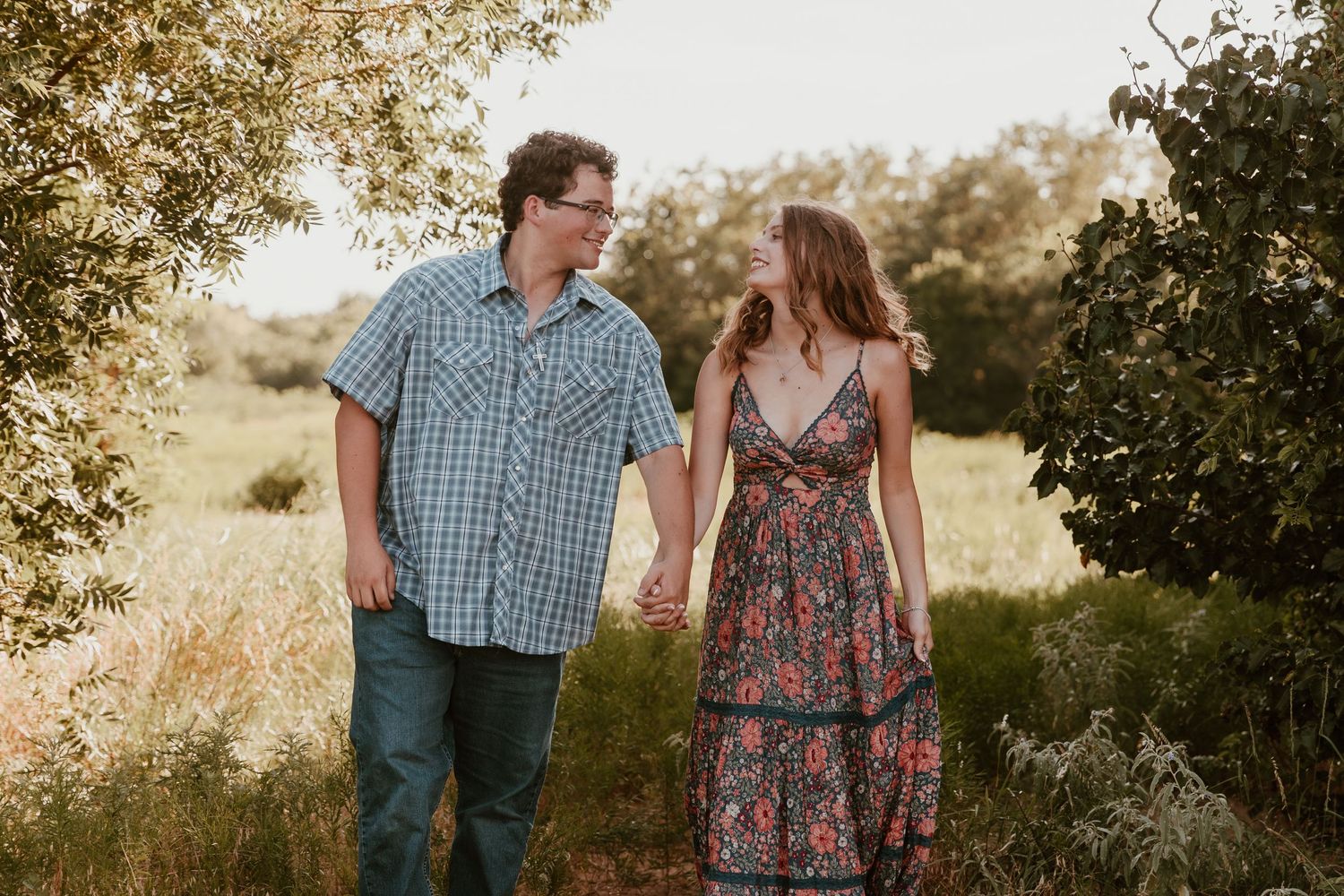 A couple walks together across a sun-drenched field during golden hour while holding hands.