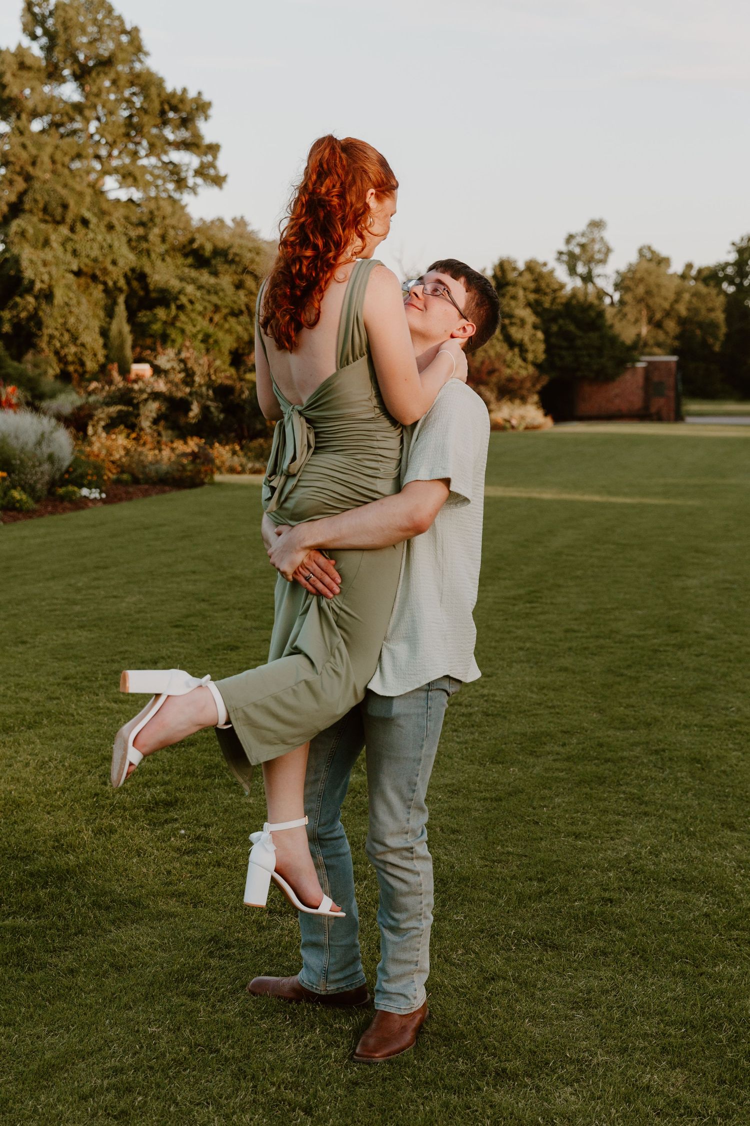 A couple shares a romantic embrace on a green lawn at sunset, with her wearing a sage dress and white heels.