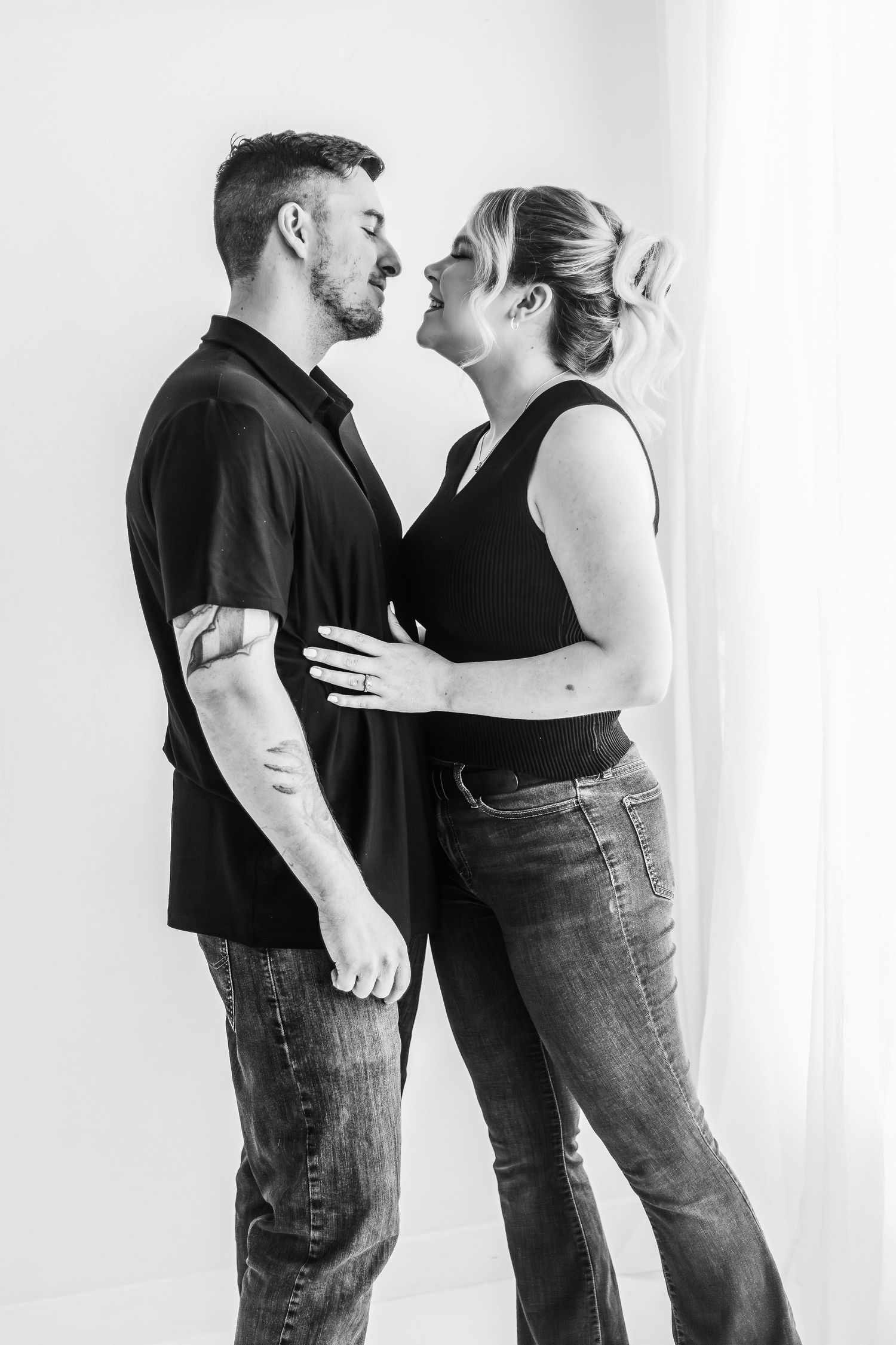 A romantic black and white portrait of a couple in casual attire sharing an intimate moment against a white wall.