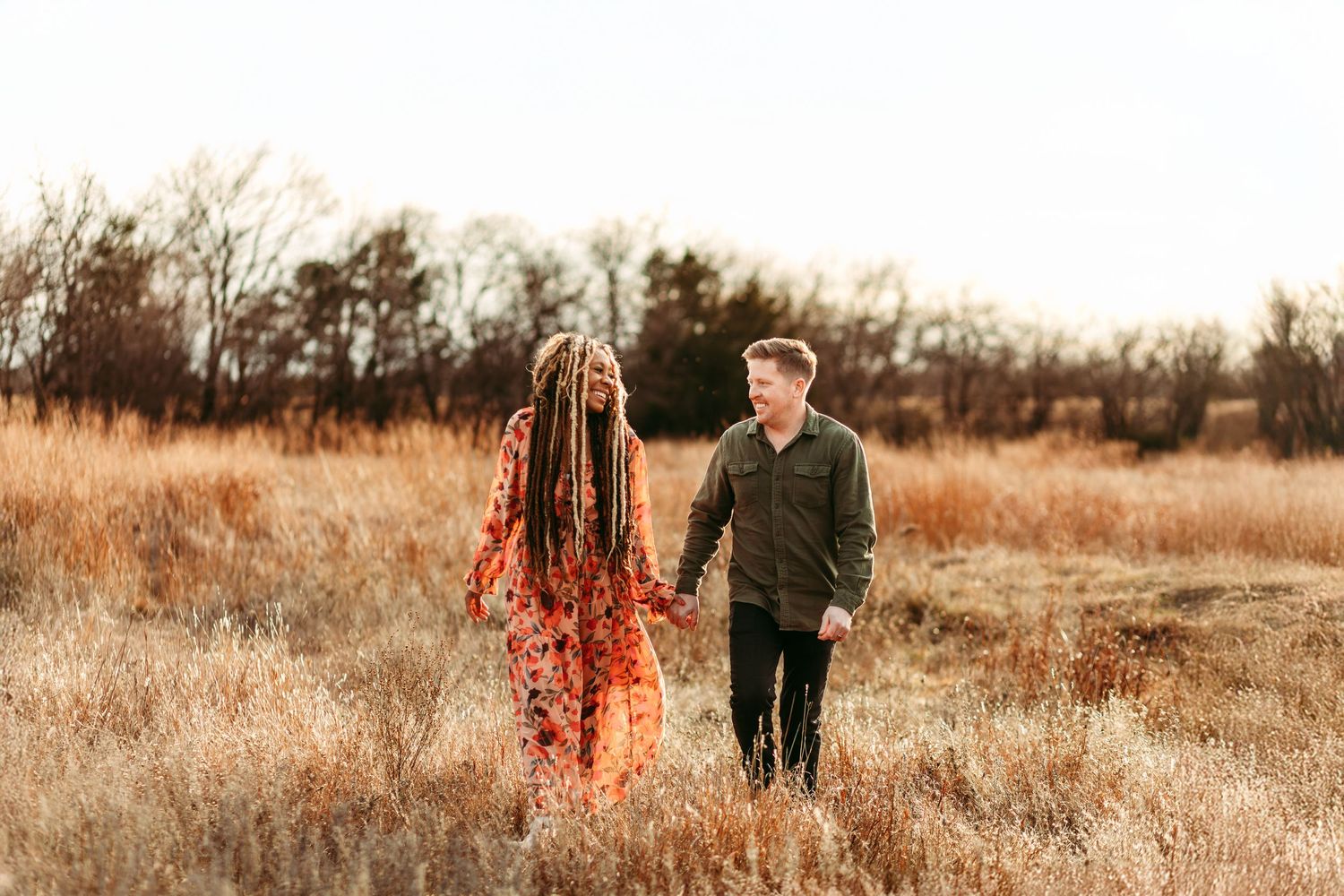 A couple stands in a golden field at sunset with one wearing a flowing red floral dress.