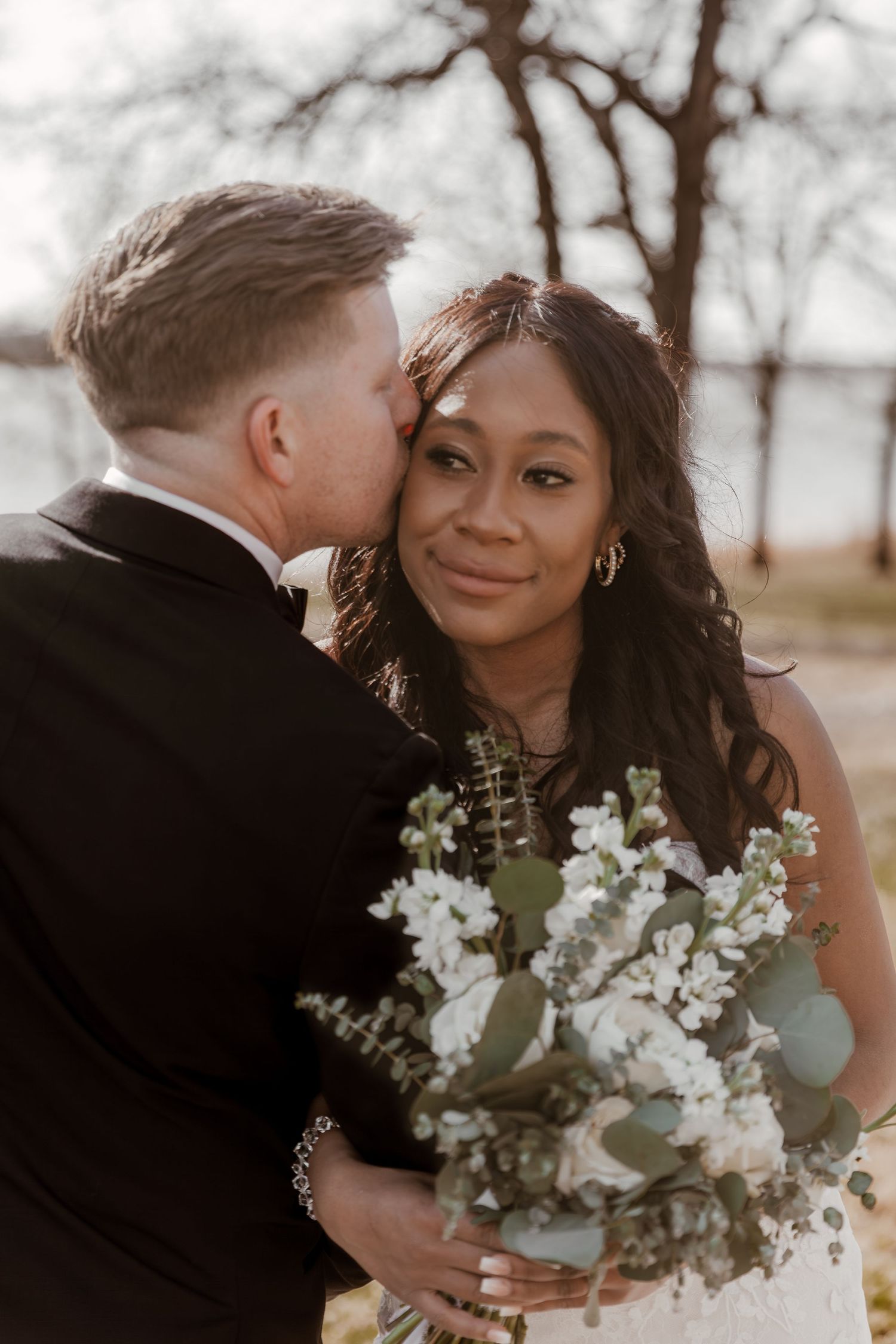 Intimate wedding portrait series showing couple embracing outdoors with white floral bouquet and winter trees in background.