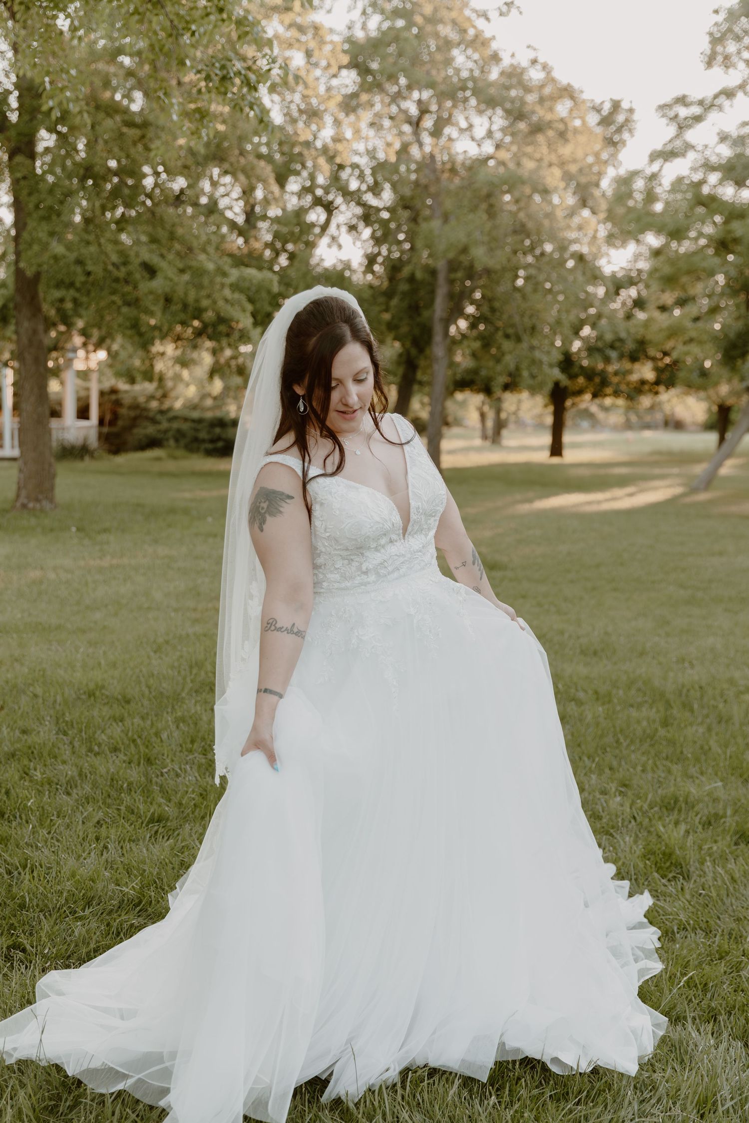 A bride in a flowing white lace wedding dress twirls playfully on a grassy field during golden hour.