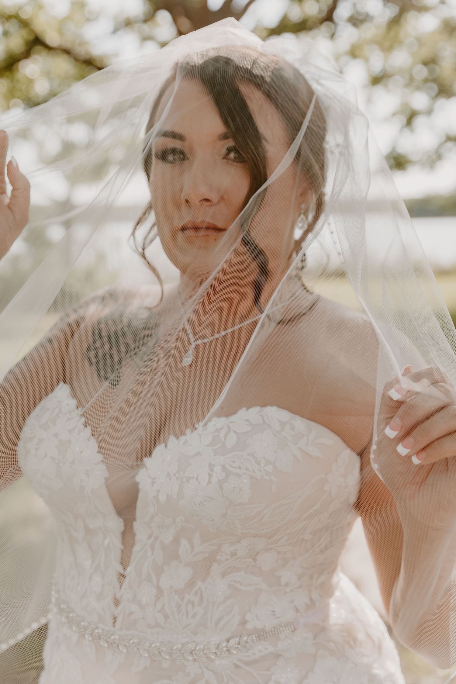 Elegant bride in a strapless lace wedding dress poses behind a sheer veil with soft bokeh background.