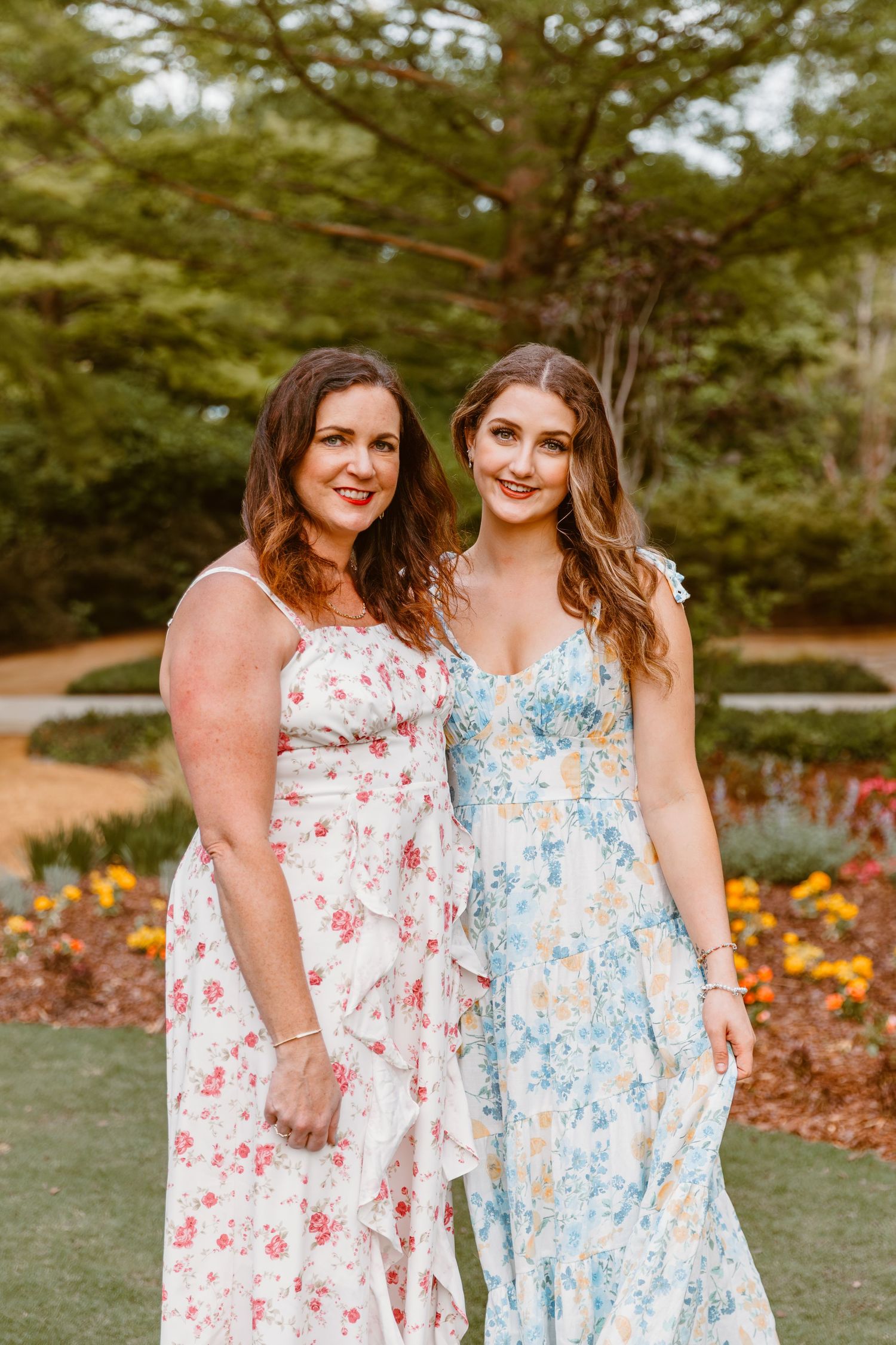 Two women pose in floral maxi dresses, one in pink and one in blue, standing in a garden with flowers and trees.