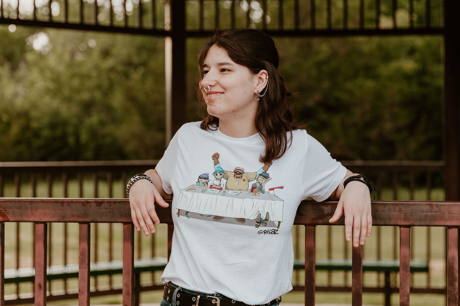 A person in a white graphic t-shirt and dark pants leans casually against a railing in an outdoor setting.