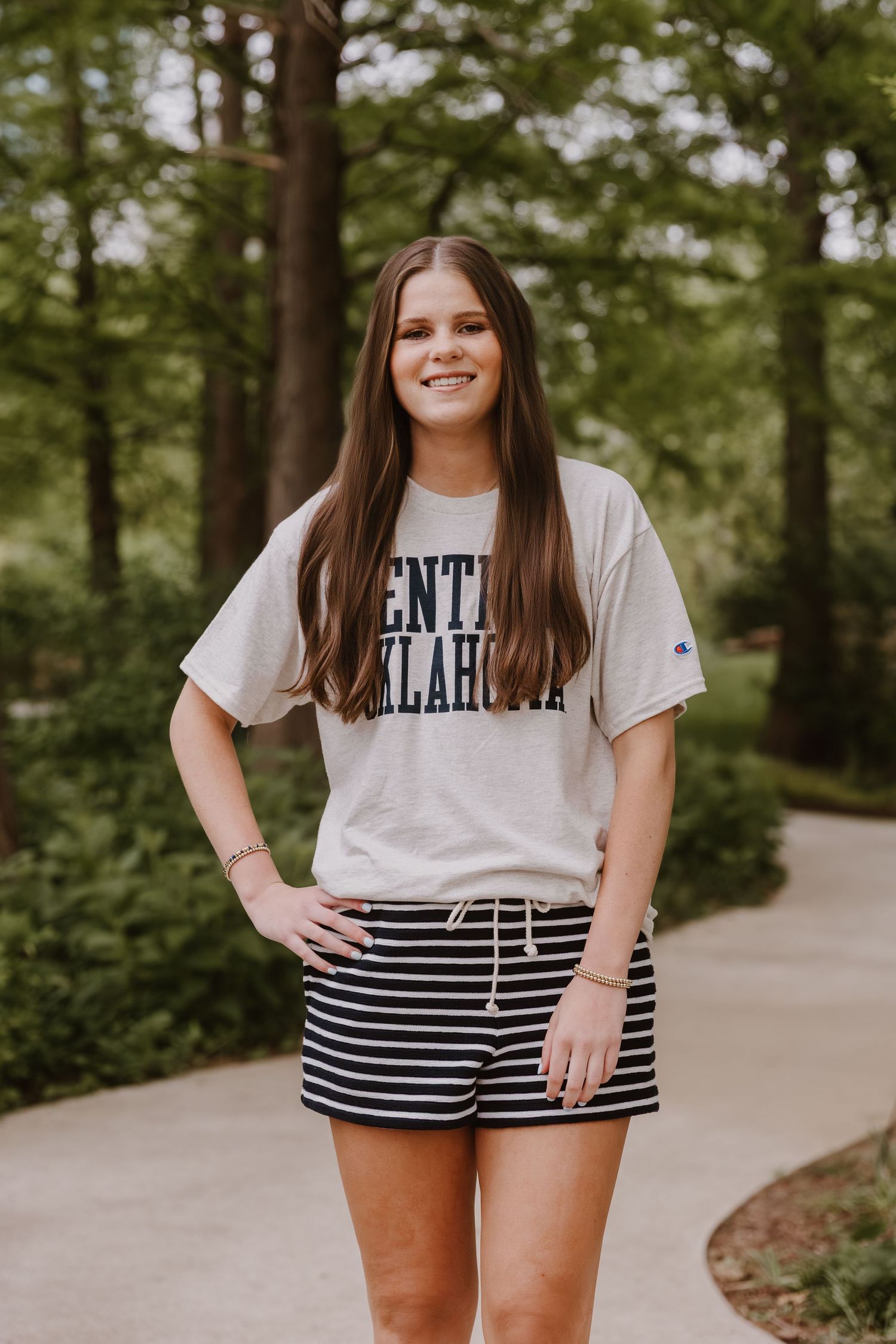 Young woman smiling in a Central Oklahoma Champion t-shirt and striped shorts, standing on a path surrounded by trees.