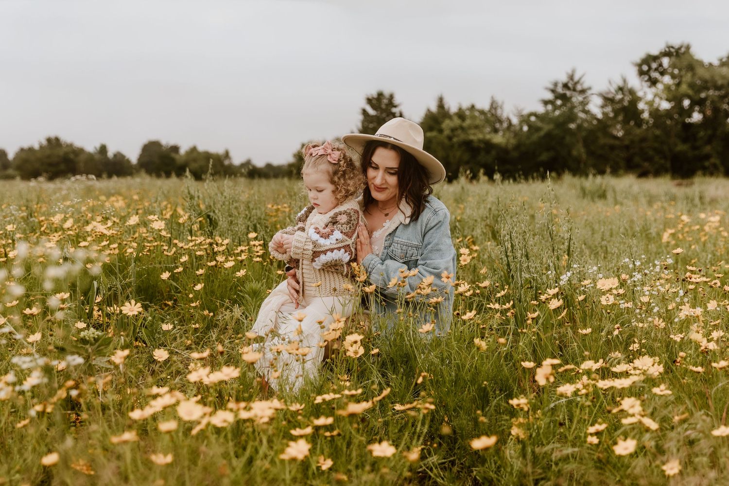 A mother and toddler sit together in a wildflower meadow, with the woman wearing a denim jacket and straw hat.