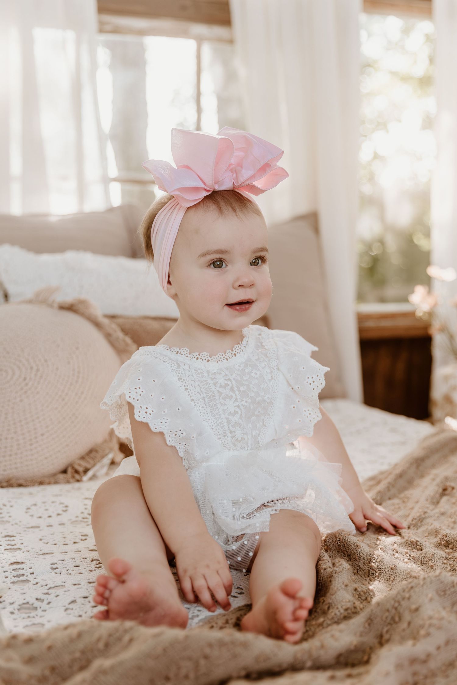 Adorable baby girl in white lace romper with pink bow headband sitting on a beige blanket in a bright, airy room.