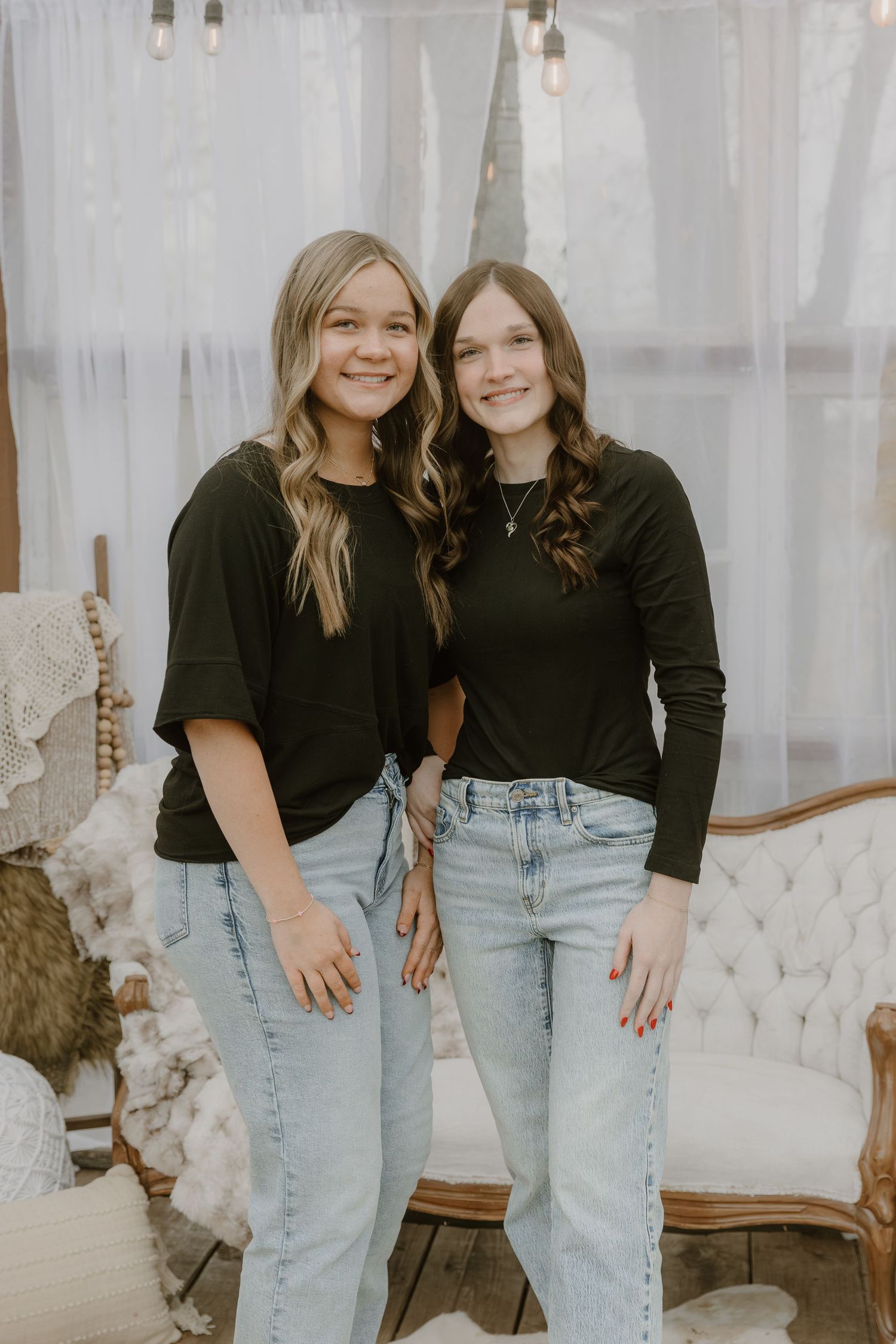 Two friends in matching black tops and light denim jeans pose together against a white curtained background.