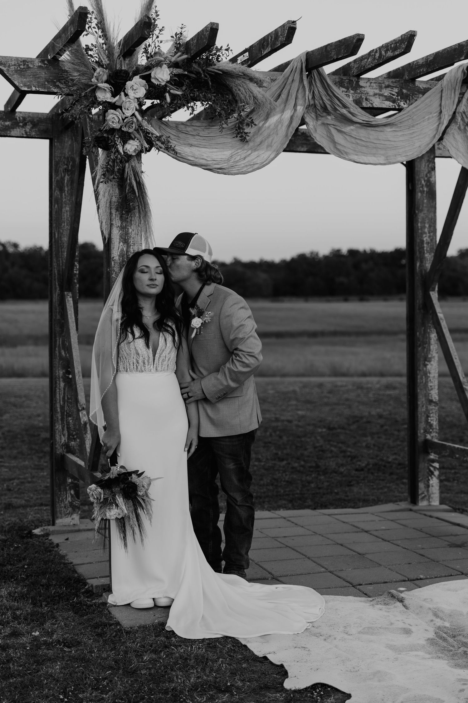 A romantic moment under a rustic wooden arch decorated with flowing fabric and floral arrangements at an outdoor wedding.