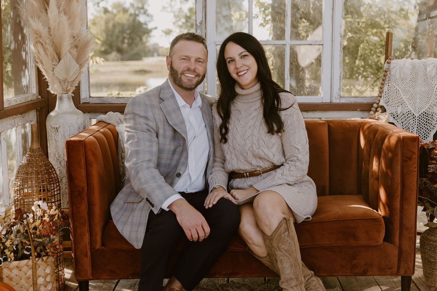 A couple sits together on a vintage brown leather couch in a sunlit room with boho decor and dried flowers.