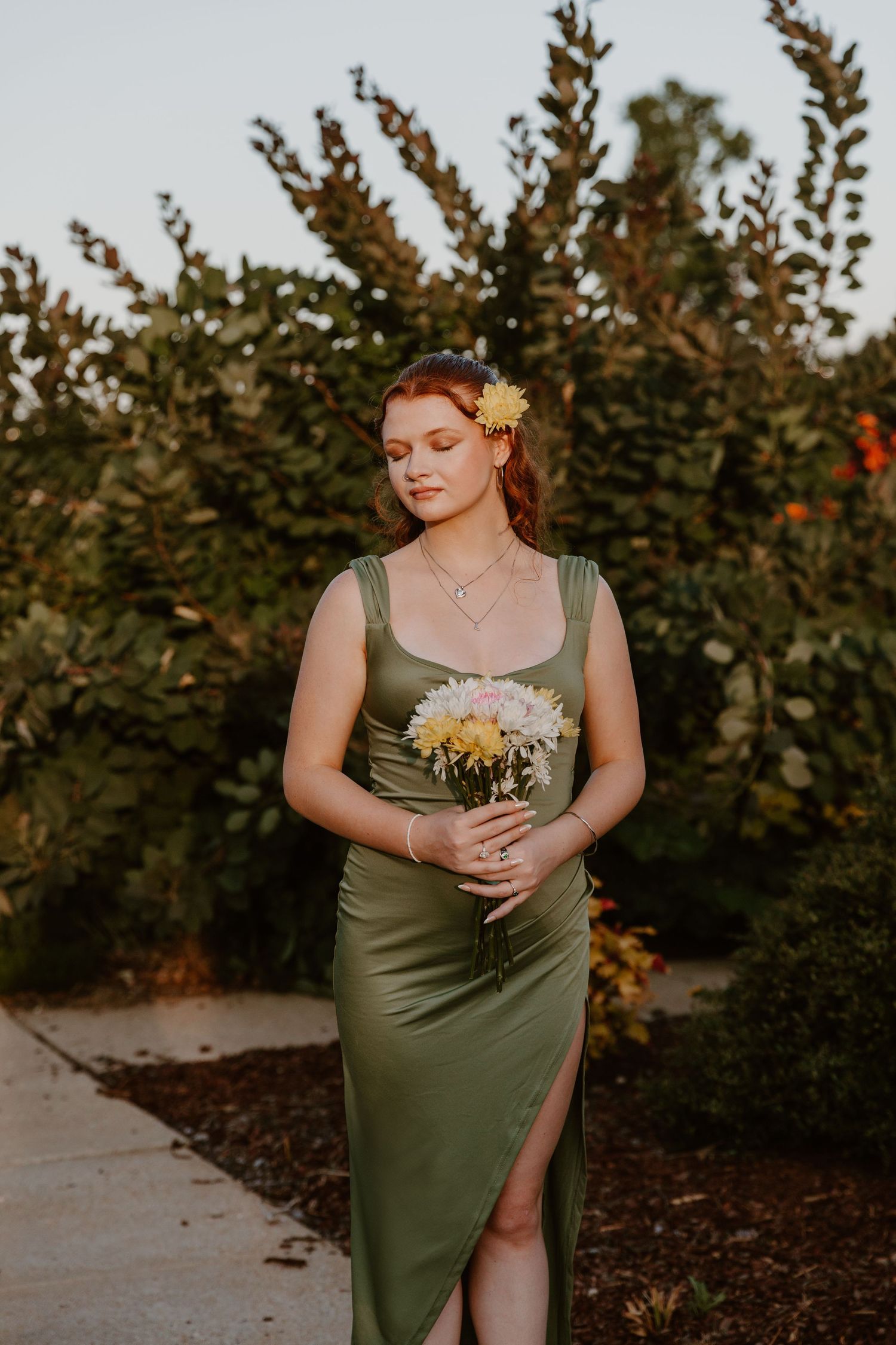 A person in an olive green dress holds a bouquet of white daisies while standing in a garden at sunset.