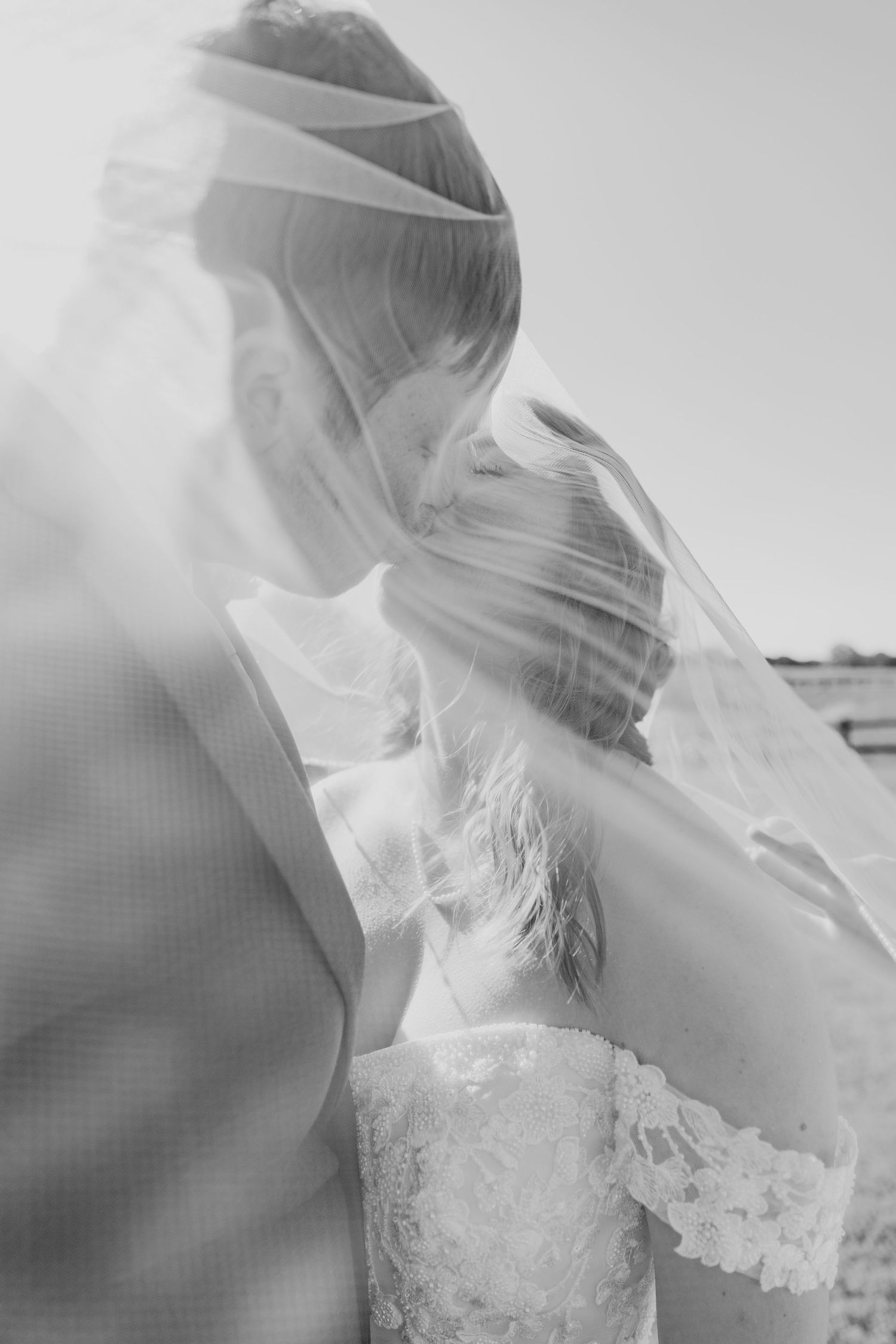 Black and white photo of bride and groom kissing under flowing wedding veil.