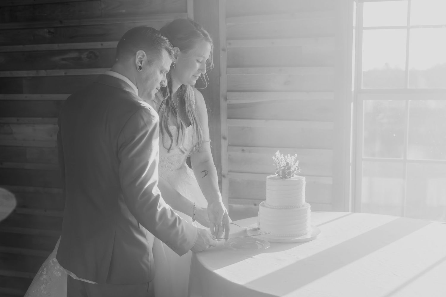 Black and white photo of a bride and groom cutting their wedding cake together in a rustic barn venue.