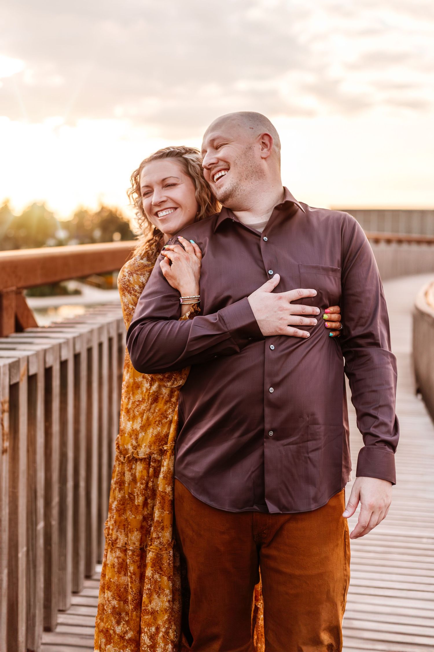 Couple sharing a tender embrace and laugh on a wooden boardwalk during golden hour sunset.