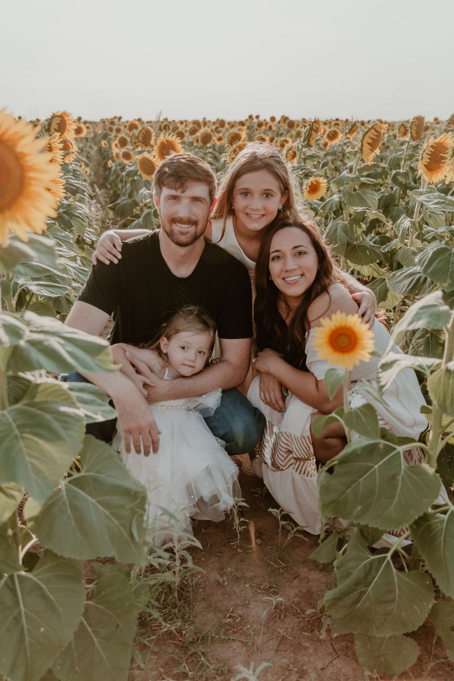 A family poses together among tall blooming sunflowers in a field during sunset.