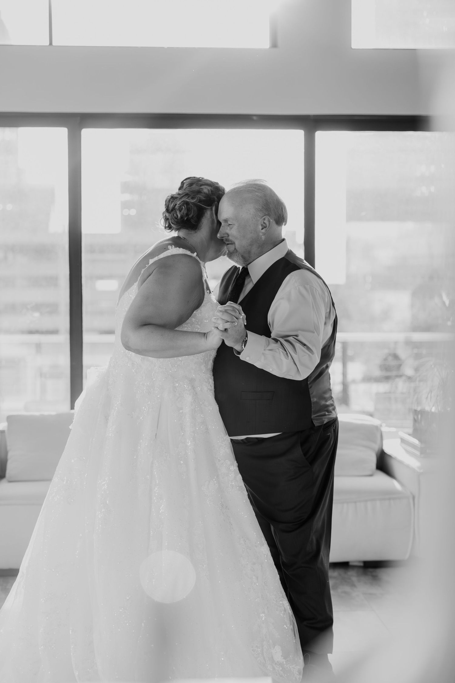 An intimate black and white wedding dance moment captured near large windows in a bright indoor venue.