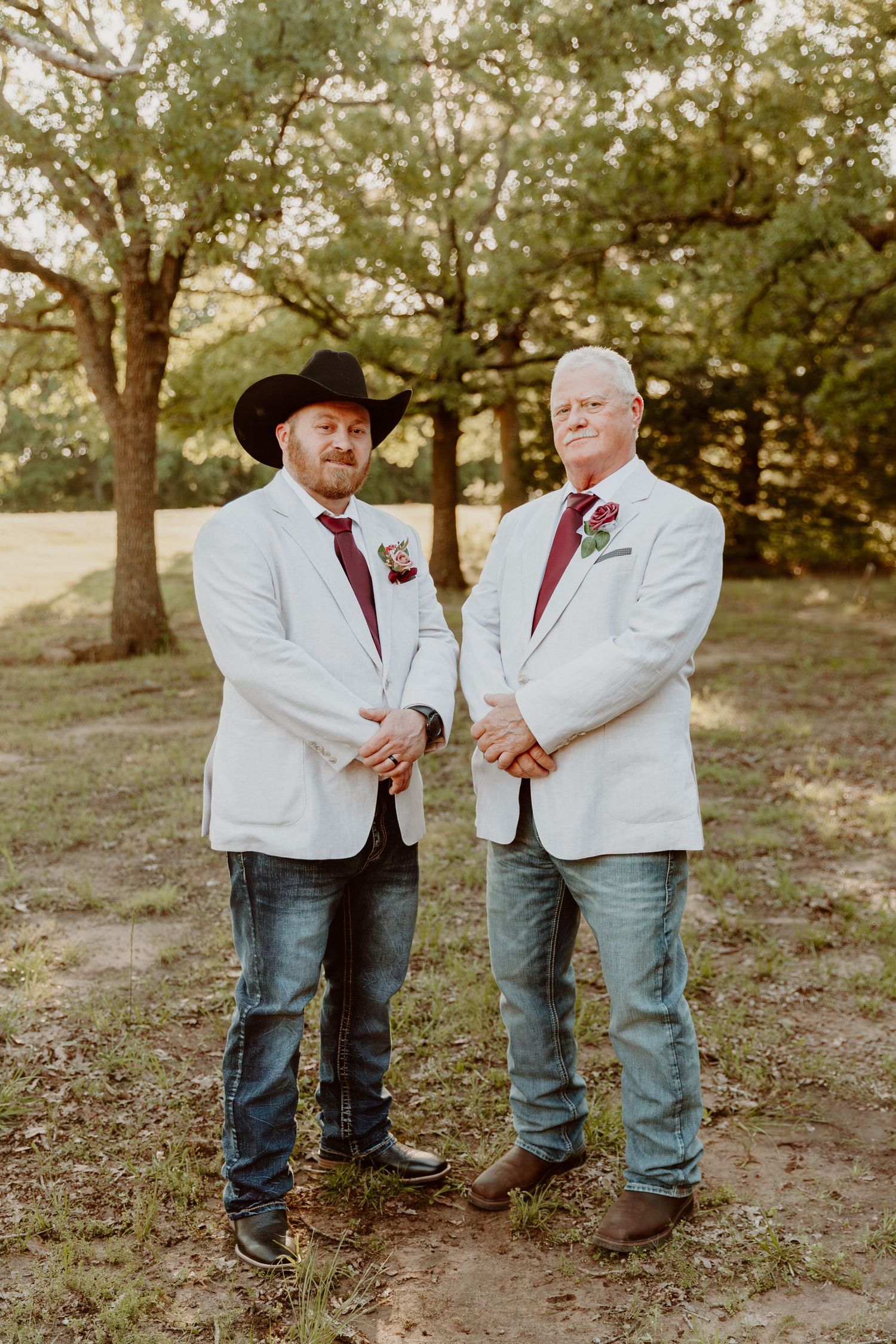Two men wear matching white jackets with red ties outdoors, one in a black cowboy hat, standing together in a wooded area.
