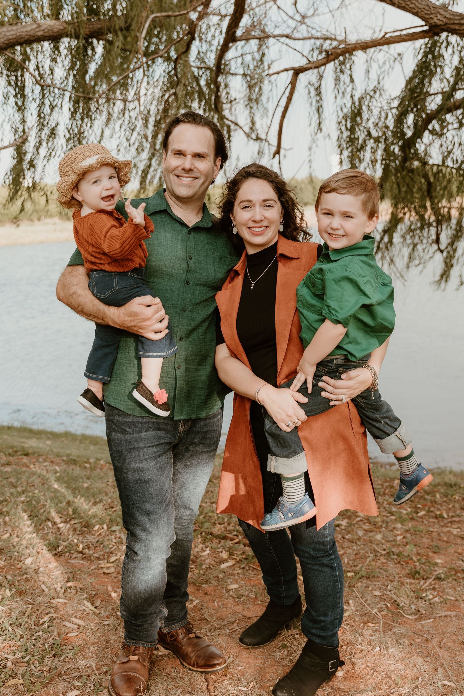 A family wearing green and orange clothing poses together outdoors near a lake on an autumn day.