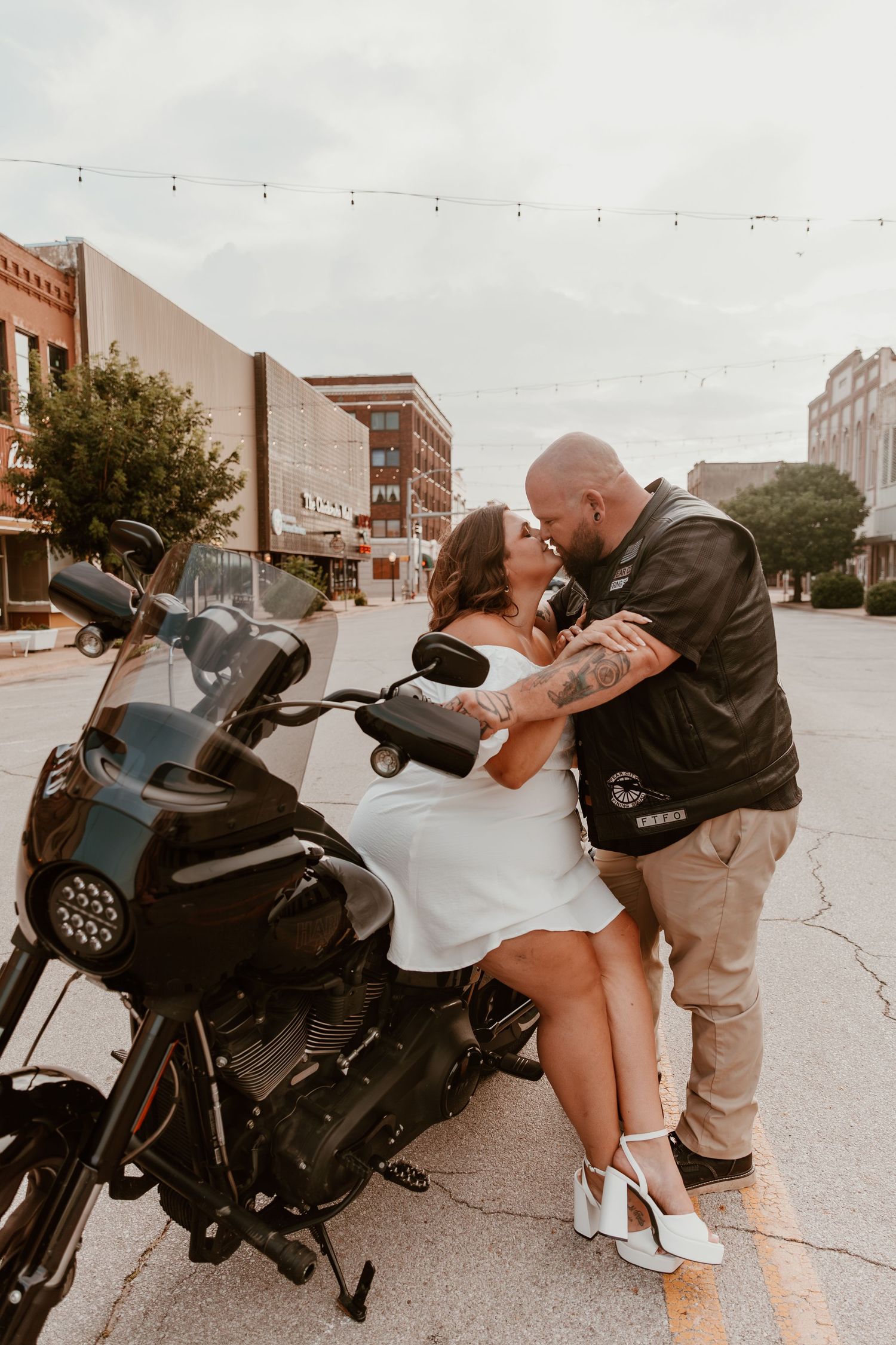 A romantic couple embraces beside a parked motorcycle on an urban street with string lights in the background.