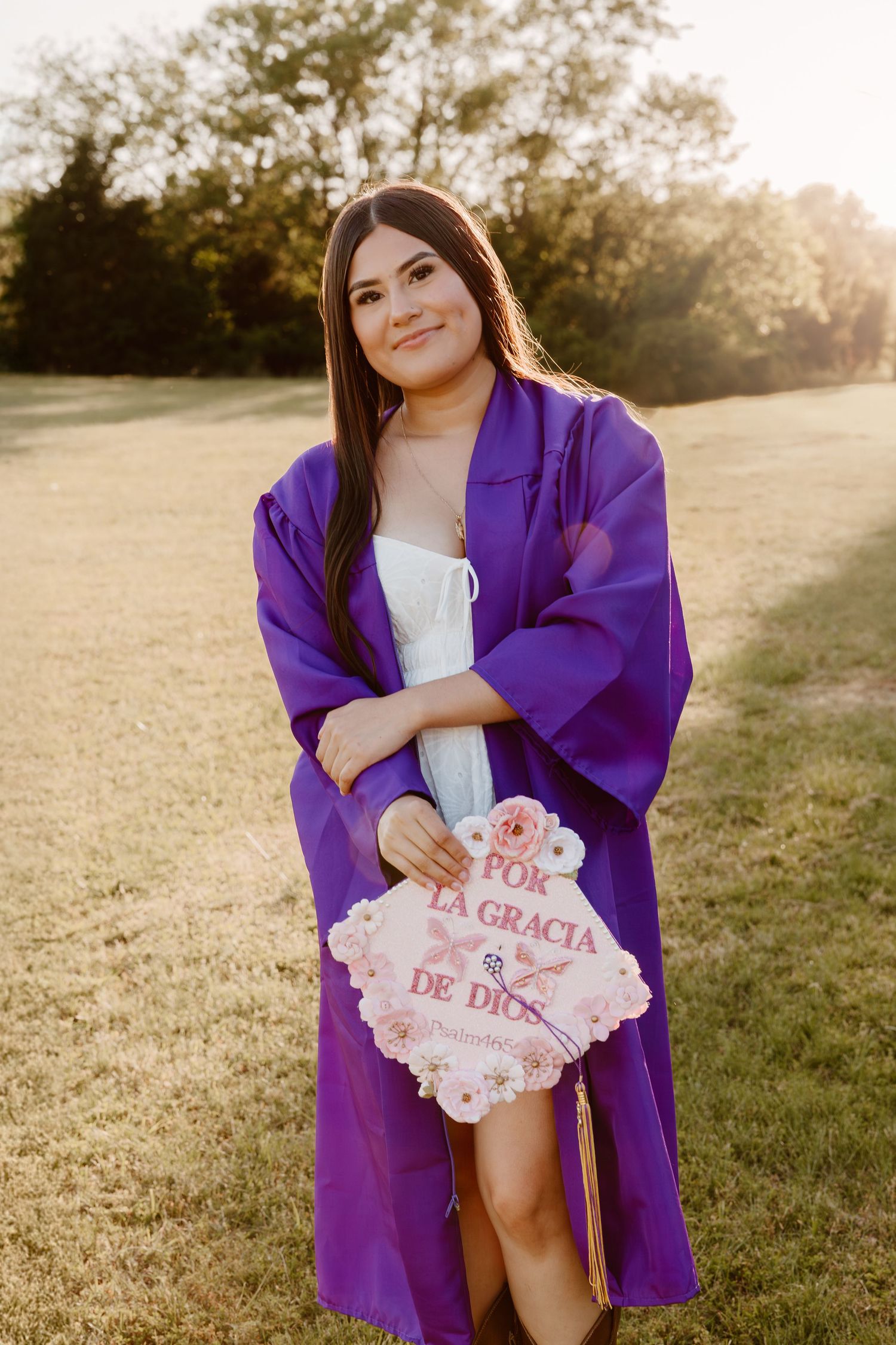 Graduate in purple graduation gown holds decorative sign while posing in sunlit field at sunset.